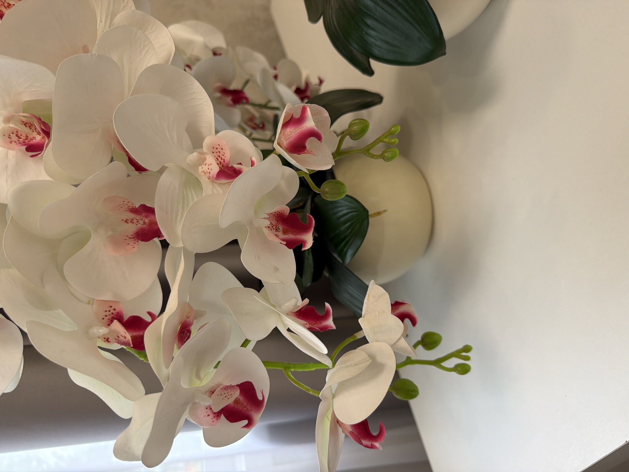 A bouquet of artificial white orchids with pink centers is displayed in a cream-colored pot on a white surface, likely a table. The orchids have delicate petals and some have small green buds. In the background, a gray curtain is visible to the left, and a light-colored wall with a textured pattern is to the right. Another artificial plant with large, dark green leaves is partially visible on the right side of the frame. The lighting is soft, suggesting an indoor setting.