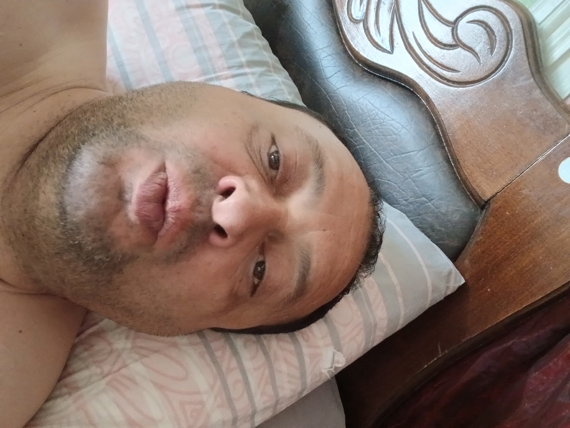 A selfie taken from a low angle, looking up at a man with a fair complexion, dark hair, and a light beard. He is lying on a pillow, with his head resting against the carved wooden headboard of a bed. The pillow is patterned with horizontal stripes in white and light gray. A portion of a dark red curtain is visible in the upper right corner of the frame. The bed appears to be in a bedroom setting. The man's mouth is slightly open, and his expression is neutral. The scene is indoors. No specific location in Telavi, Georgia is identifiable.