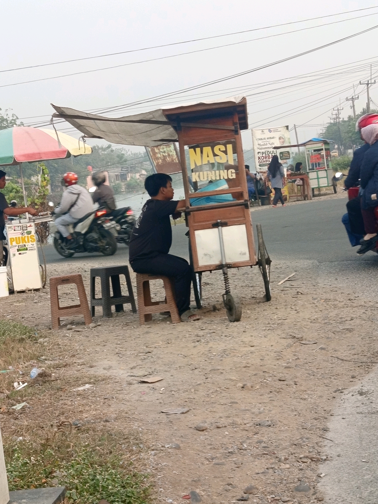 A vendor is sitting on a stool beside a food cart that says "NASI KUNING". The cart is on a dirt road. There are other food carts in the background, as well as people and motorcycles. The sky is overcast. The scene appears to be in a market or roadside area in Karawang, Indonesia.