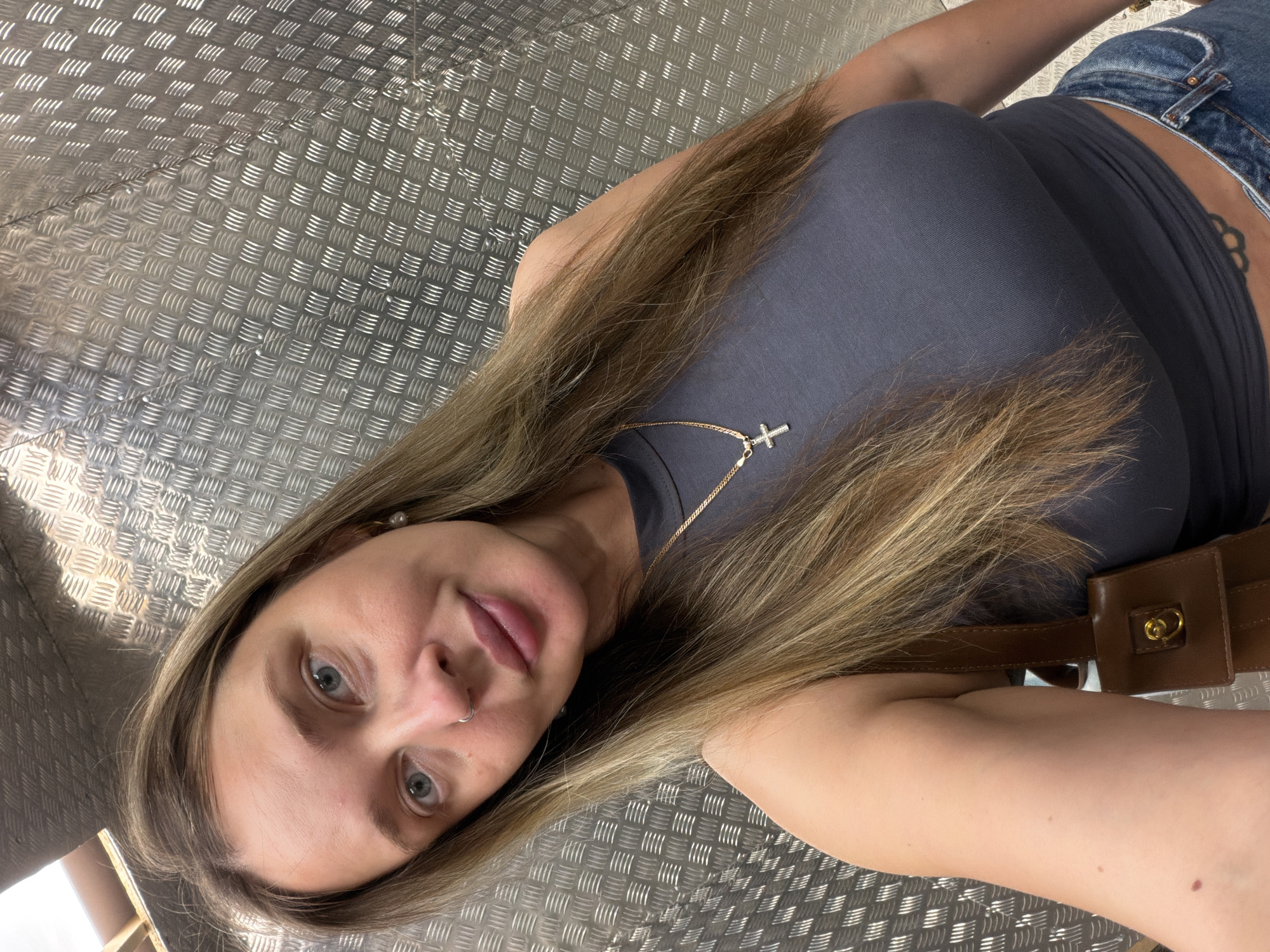A woman with long, blonde hair, wearing a dark gray tank top and blue jeans, poses for a selfie in a metallic-walled room. She has a nose ring and a gold chain with a cross pendant. A brown purse is visible on her left side. The background appears to be a textured metal surface, possibly the interior of an elevator or a service area. There is no indication of her location in Moscow, Russia.