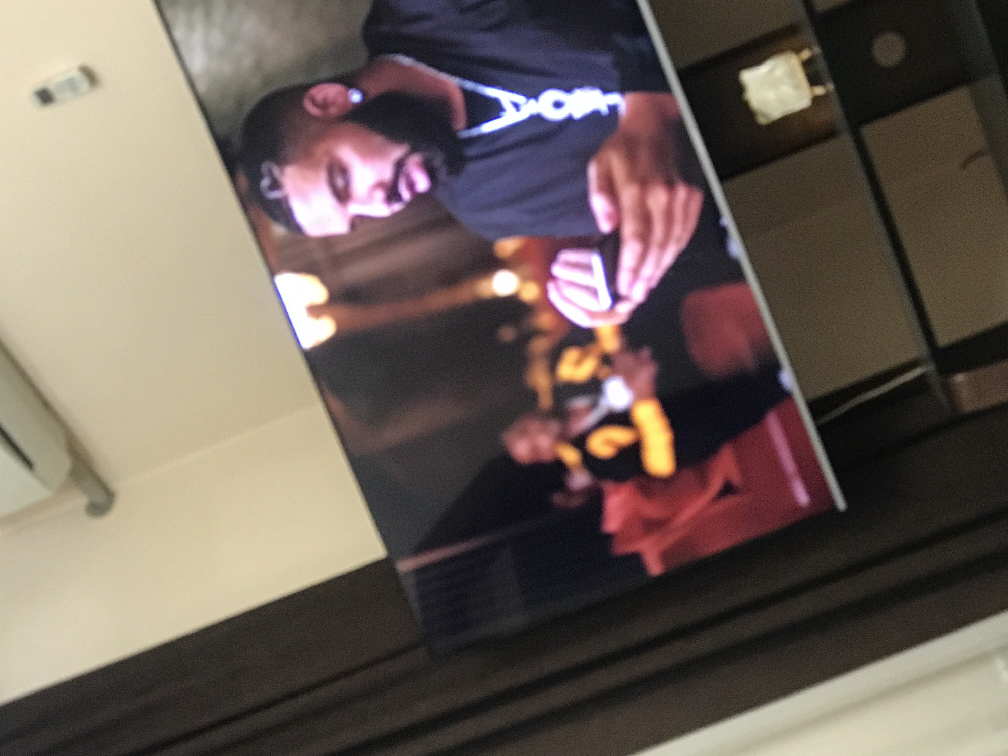 The image depicts a television screen displaying a scene with two individuals. One person in the foreground wears a black shirt with white lettering and appears to be indoors with soft lighting. In the background, another individual wears a yellow and black jersey, likely in an outdoor setting. The television is mounted on a wall above a dark brown surface. There is a white object to the left of the television on the wall. The environment appears to be an indoor setting, possibly a residential or office space.
It is not possible to determine if this scene is in Yerevan, Armenia based on this image.