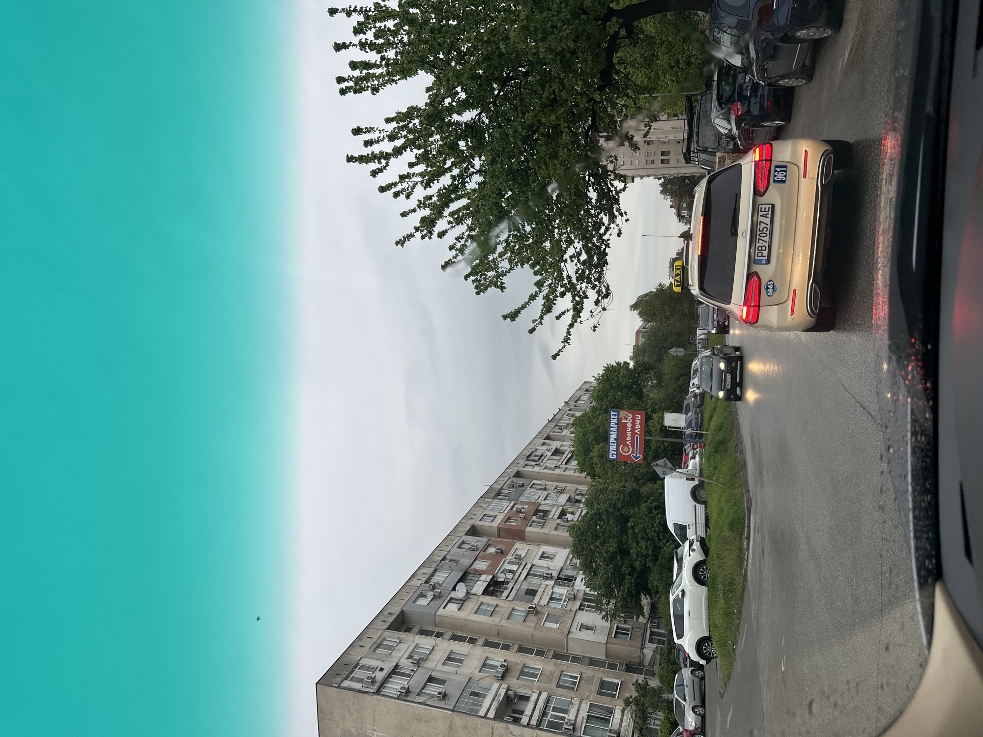The image captures a street scene in Plovdiv, Bulgaria, on what appears to be a cloudy or rainy day. A multi-story residential building with numerous windows and air conditioning units is visible along the street. A sign for a supermarket, with the words "СУПЕРМАРКЕТ Слънчеви Лъчи" (Supermarket Sunny Rays), points downwards and appears to be advertising the direction to the market. Cars are parked along the street, and traffic is congested, with a beige Mercedes station wagon taxi with license plate "PB 7057 AE" visible in the foreground, its brake lights illuminated. There's a tree with green foliage partially obscuring the view. The sky is overcast and appears grey. The overall impression is that of a typical residential street in an urban area.