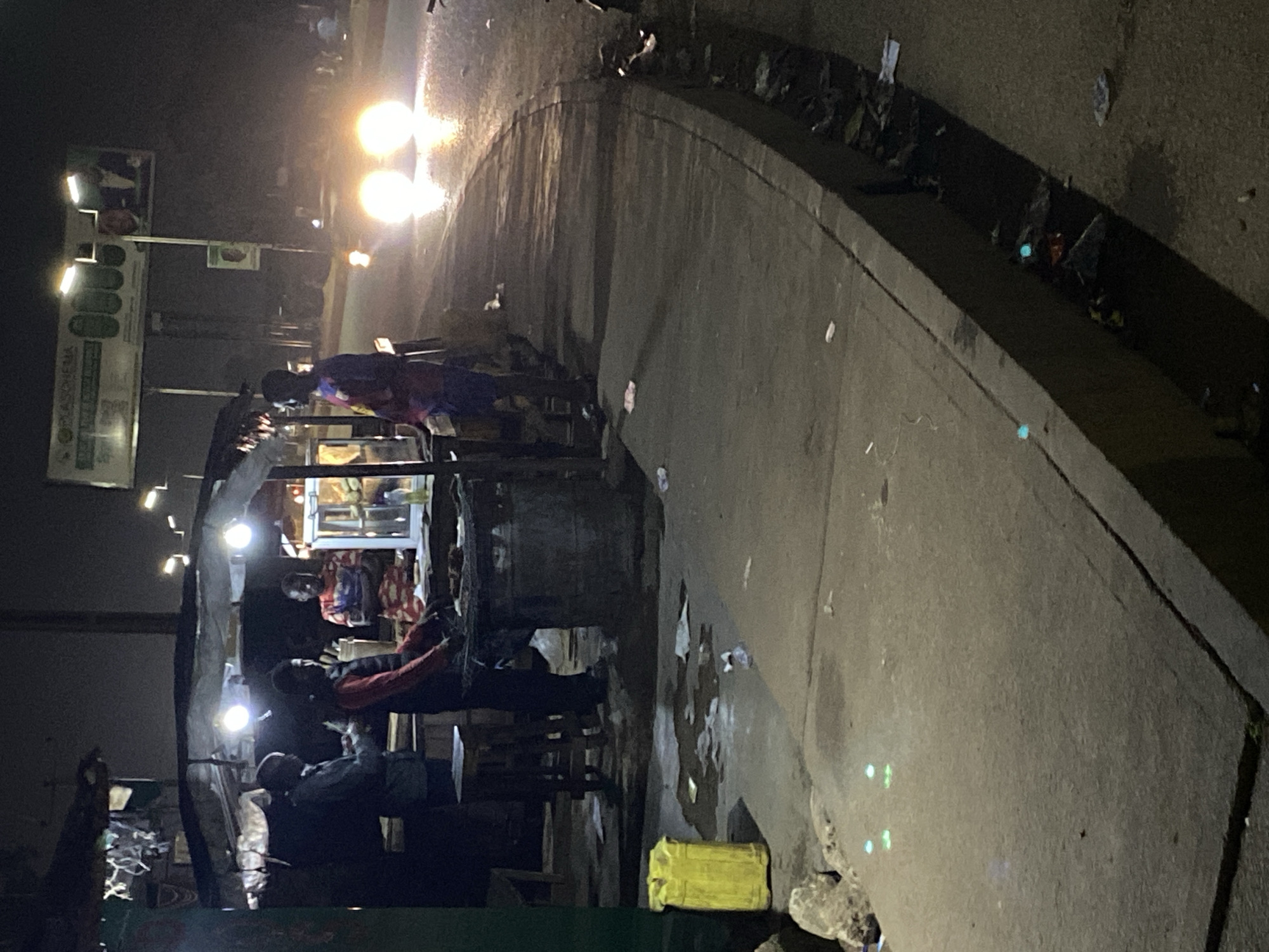 This image depicts a street scene at night in Bukuru, Nigeria, focused on a roadside food stall. Three individuals are visible, two actively engaged with the food stall and one standing to the side. One person, wearing a red vest over a dark shirt, appears to be preparing food on a grill or hot surface. Another person in a patterned shirt stands behind the counter, and a third person in a dark jacket is on the far left, possibly a customer. The food stall is illuminated by bright overhead lights, creating a focal point. A yellow container is present on the sidewalk to the left of the stall. The street is wet, reflecting the lights from passing vehicles, two of which have their headlights on. The sidewalk is littered with debris, and a drainage ditch alongside the sidewalk also contains trash. A large billboard with text and images is visible in the background.
