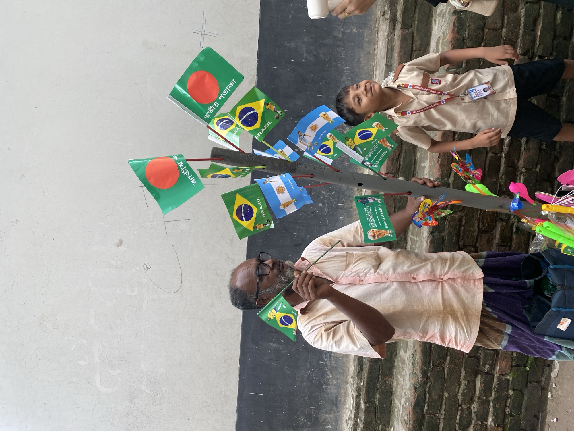 A street vendor stands in front of a rough, white, cracked wall in an urban environment. He is a man with dark skin, short grey hair, a beard, and glasses. He wears a light pink button-down shirt and a patterned wrap skirt. He holds a small Brazilian flag in his right hand. To his right, a young boy in a tan school uniform with a red lanyard stands looking up and to the right. A collection of flags, including those of Brazil, Argentina, and Saudi Arabia, are displayed on a vertical stand behind the vendor and boy. The ground is made of brick and rough dirt.
