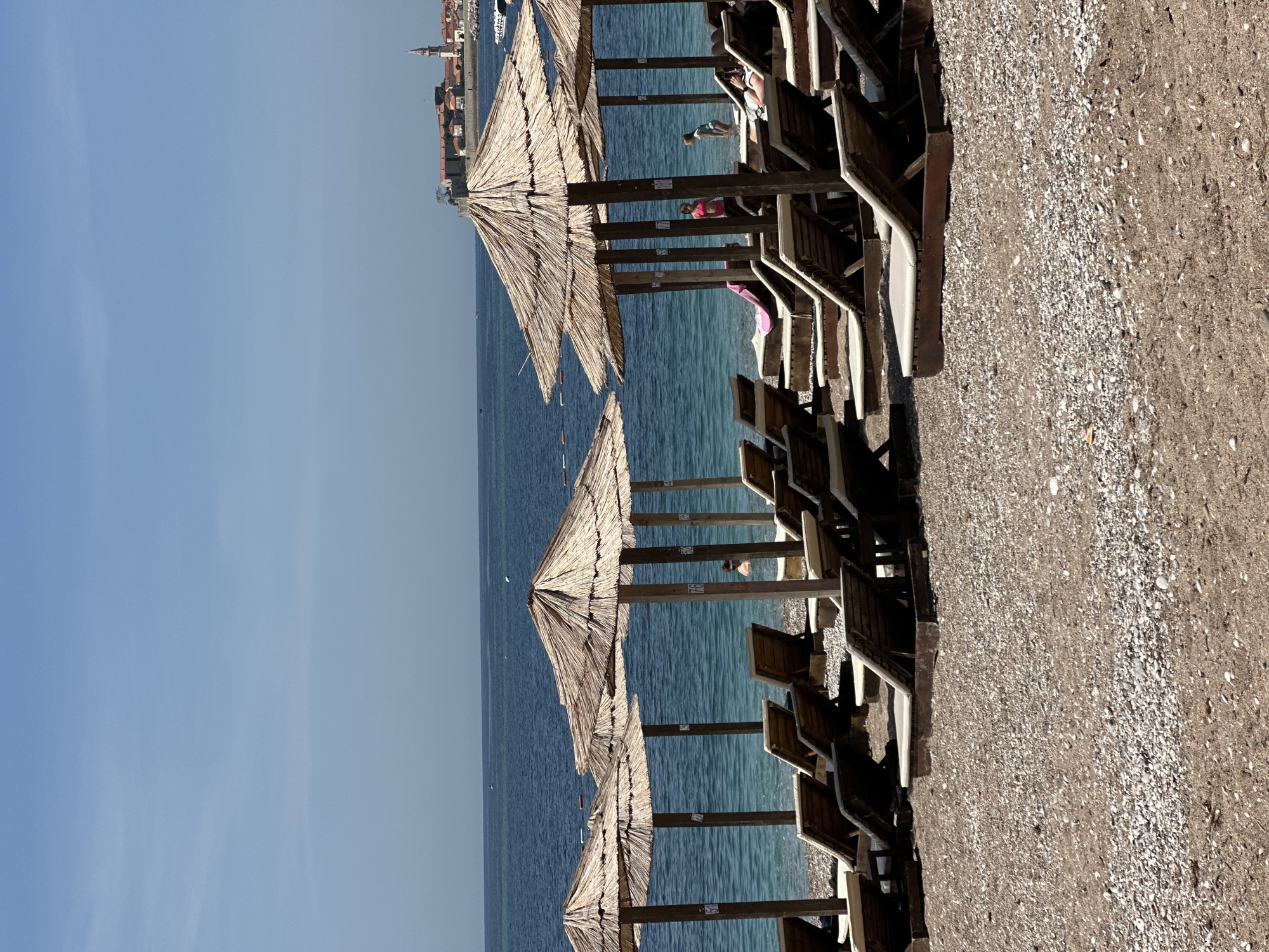 The image shows a beach scene in Budva, Montenegro. The scene consists of rows of thatched umbrellas and wooden lounge chairs on a pebbly beach. The Adriatic Sea is visible behind the umbrellas, with a view of the old town of Budva in the distance. People are partially visible between the umbrellas, appearing to be swimming or relaxing in the water. The sky is clear and blue.