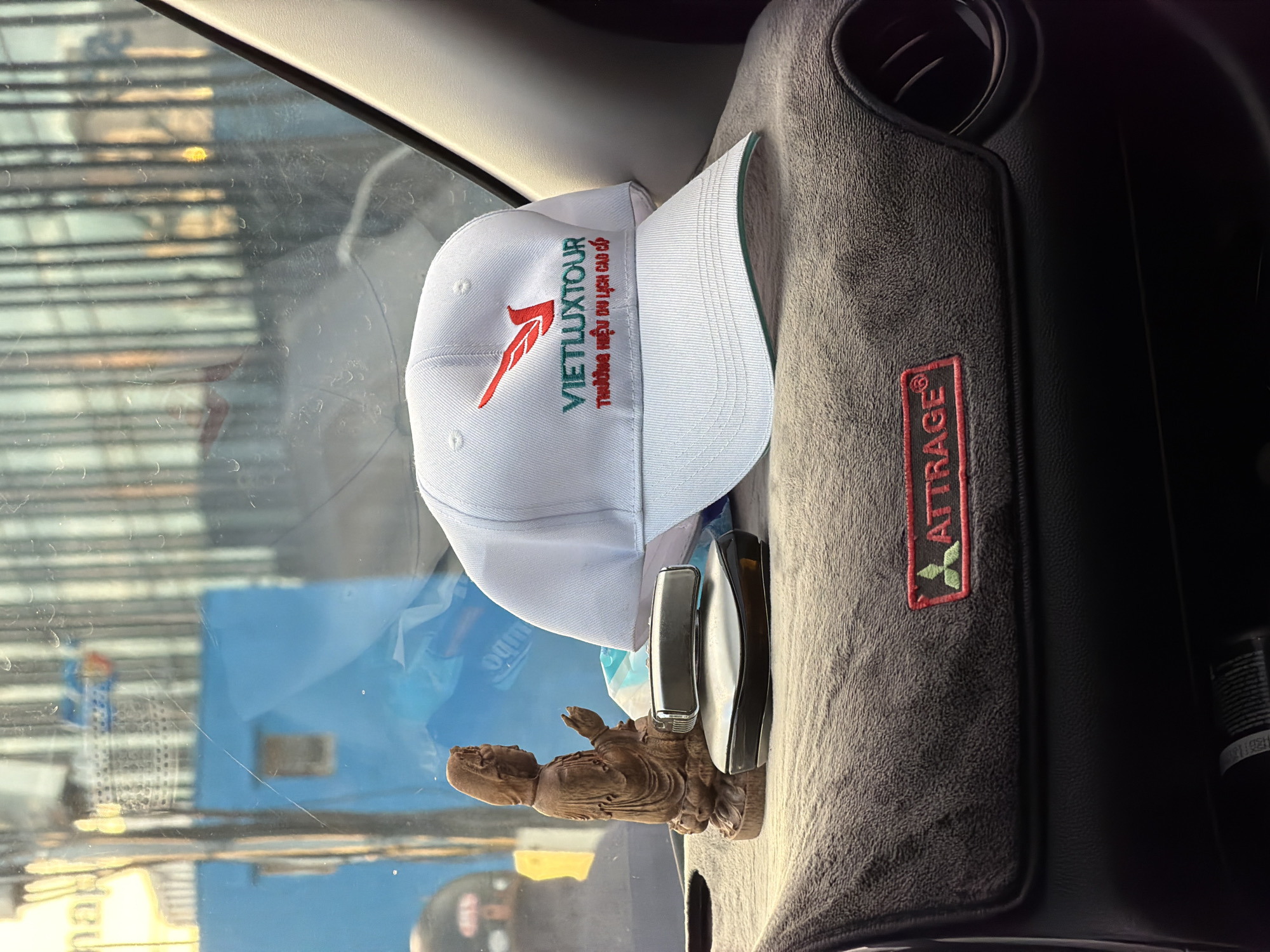 The image depicts a close-up view of a car's dashboard in what appears to be Đà Nẵng, Vietnam. The primary focus is a white baseball cap with red and green embroidery, displaying the text "VIETLUXTOUR" and a smaller Vietnamese phrase below. To the left of the cap is a small wooden figurine, possibly a deity, and a black electronic device with a metallic strip. The dashboard itself is covered with a gray fuzzy mat, featuring a red rectangular patch with green embroidery of a tree-like symbol and the word "ATTRAGE". In the background, visible through the car's windshield, are buildings and structures that suggest an urban environment, with reflections indicating it is daytime. The overall scene is static and appears to be taken from inside the vehicle.