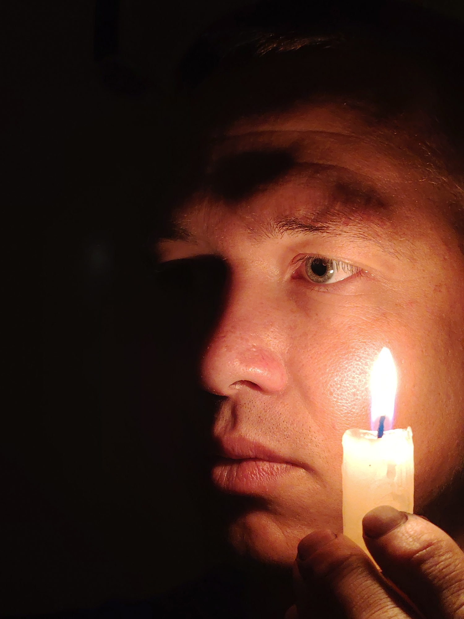 A close-up, eye-level, medium shot captures a person's face in the dark, illuminated by a single lit candle held in their hand. The subject's right eye is visible, with a light brown iris and a slight reflection from the flame. The candlelight casts a warm glow on the right side of their face, highlighting textures of the skin and creating deep shadows on the left. The candle itself, a thick white pillar with a blue wick, is held in a hand that is partially visible in the foreground. The background is almost entirely black, providing no environmental context or geographical information. Therefore, the location in Chaikovsky, Russia cannot be determined from the image.