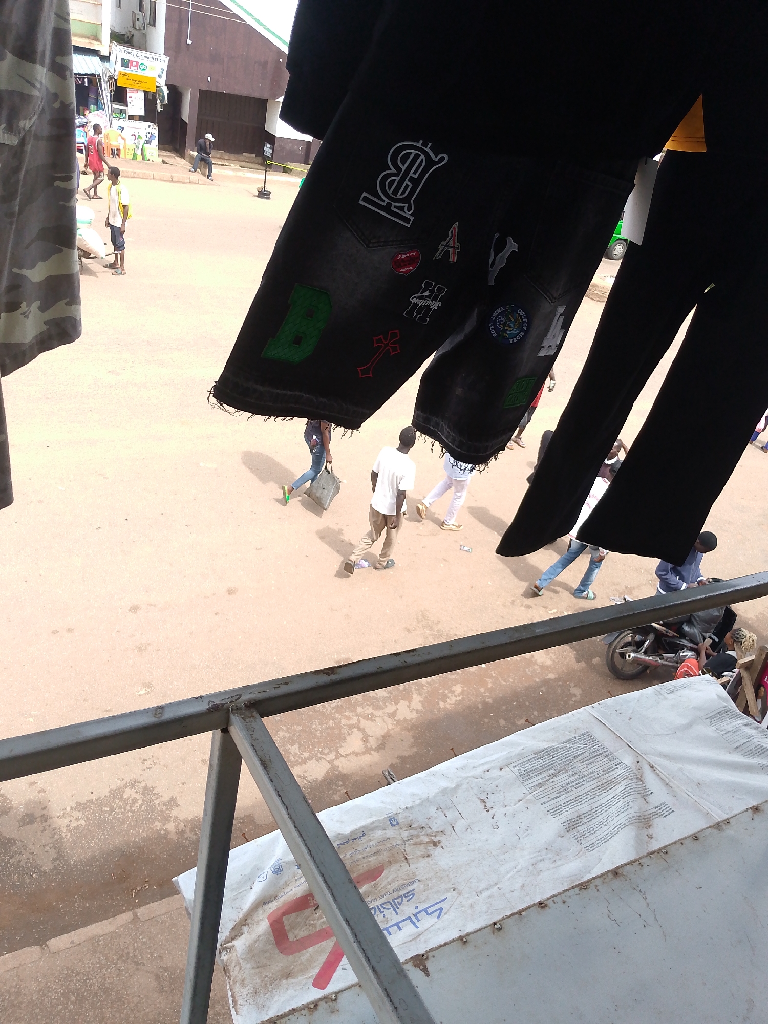 The image captures a street scene in Bukuru, Nigeria, with a primary focus on clothing displayed for sale. In the foreground, dark-colored pants with various patches, including the letters "B" and "A", are hung prominently. The background shows a street with several people walking and interacting. One person, wearing a white t-shirt and khaki pants, walks away from the camera. Another person in a dark blue jacket is seen near a motorcycle. The environment appears to be an outdoor market or commercial area, with buildings and a road visible. The ground is dusty, and there are signs of everyday life, such as a parked motorcycle and people engaged in their daily activities.