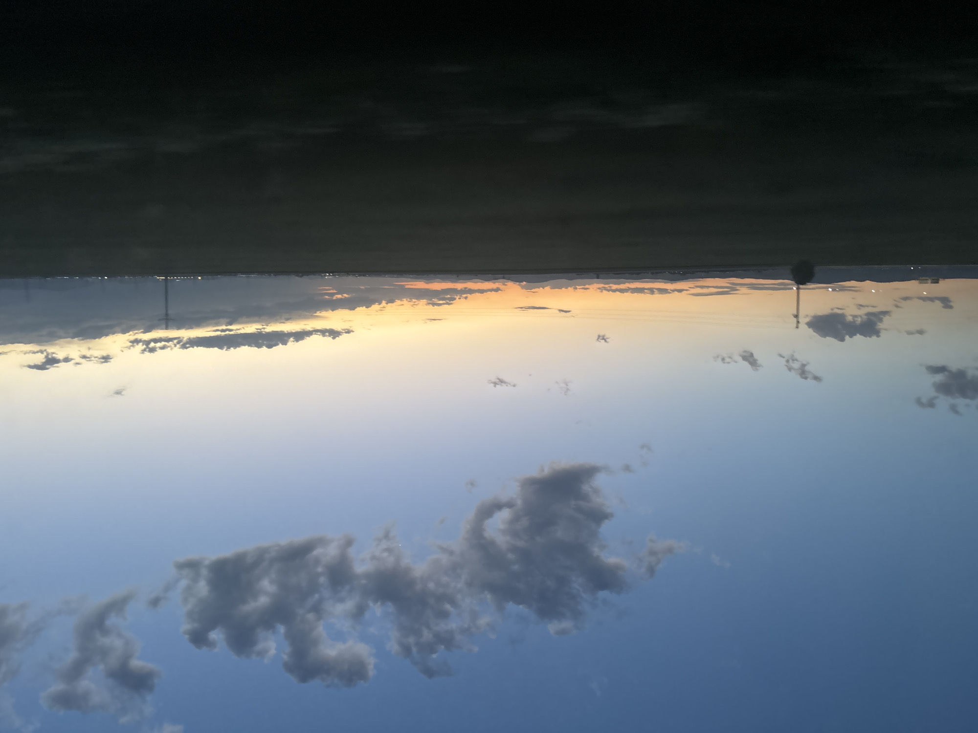 The image shows a low-angle view of the sky at dusk or dawn. The lower portion is dominated by a blue sky with scattered gray clouds. Higher up, the sky fades to a pale yellow and orange horizon, suggesting the position of the sun. On the horizon, there are faint lights suggesting a distant cityscape. Two tall structures, potentially cell towers or utility poles, are visible, piercing the horizon. The top of the image shows what appears to be the edge of a structure or roof, casting a dark shadow.
Given the prompt indicating the location as Mramor, Bulgaria, the faint cityscape on the horizon could be a distant view of Sofia or another nearby town. The tall structures likely support communication infrastructure in the area.
