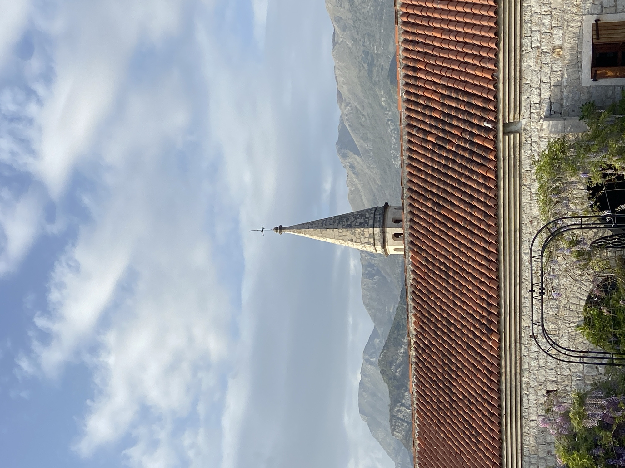 The image shows a church spire rising into a cloudy sky in Budva, Montenegro. Mountains form a hazy backdrop behind the spire. A tiled roof runs along the right edge of the frame, with a stone wall and iron archway adorned with greenery and purple flowers. A partially obscured window is visible in the top right corner of the stone wall.