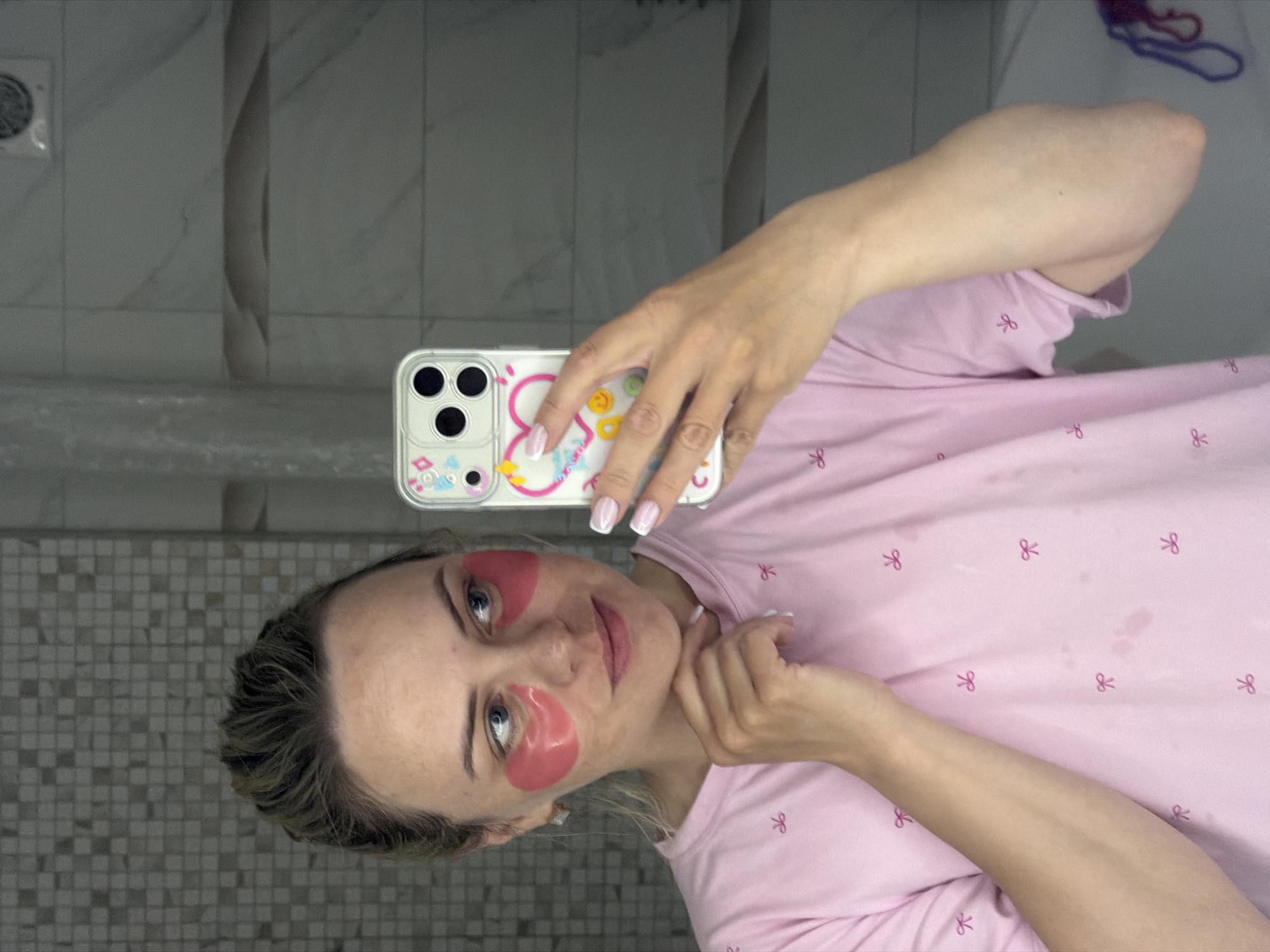 The image depicts a woman in what appears to be a bathroom. She is wearing pink under-eye patches and a light pink T-shirt with a bow pattern. She is holding up a smartphone with a clear, decorated case, seemingly taking a selfie in front of a mirror. The bathroom features gray and white tiles on the wall. A ventilation grille is visible in the upper left corner. The overall orientation of the image suggests it may have been taken in a reflective surface, possibly a mirror, thus the rotated appearance. There is no information to determine the location in the city of Votkinsk, Russia.