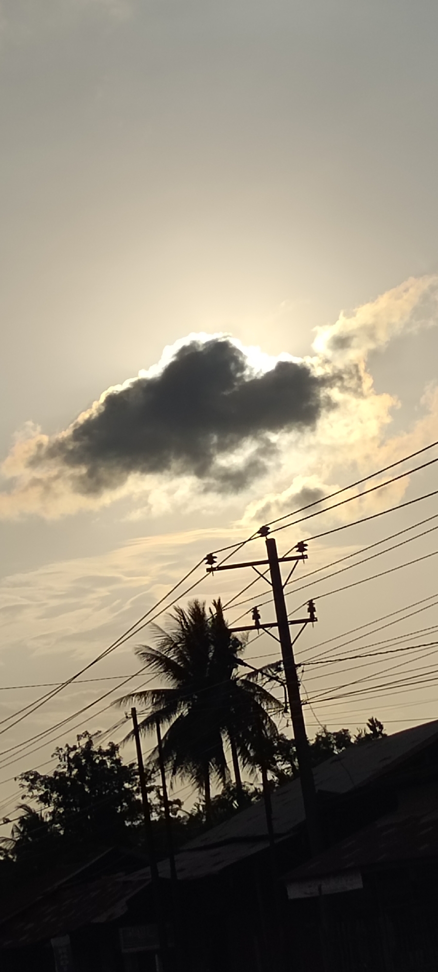 The image, likely taken in Keudeteupinpunti, Indonesia, shows a low-angle view against a pale sky with a dark cloud partially obscuring the sun. A silhouetted coconut palm tree and utility pole with power lines dominate the middle ground. Other trees and a rooftop structure are silhouetted in the foreground, suggesting an urban or suburban setting. No people are visible. The scene appears to be near dusk.