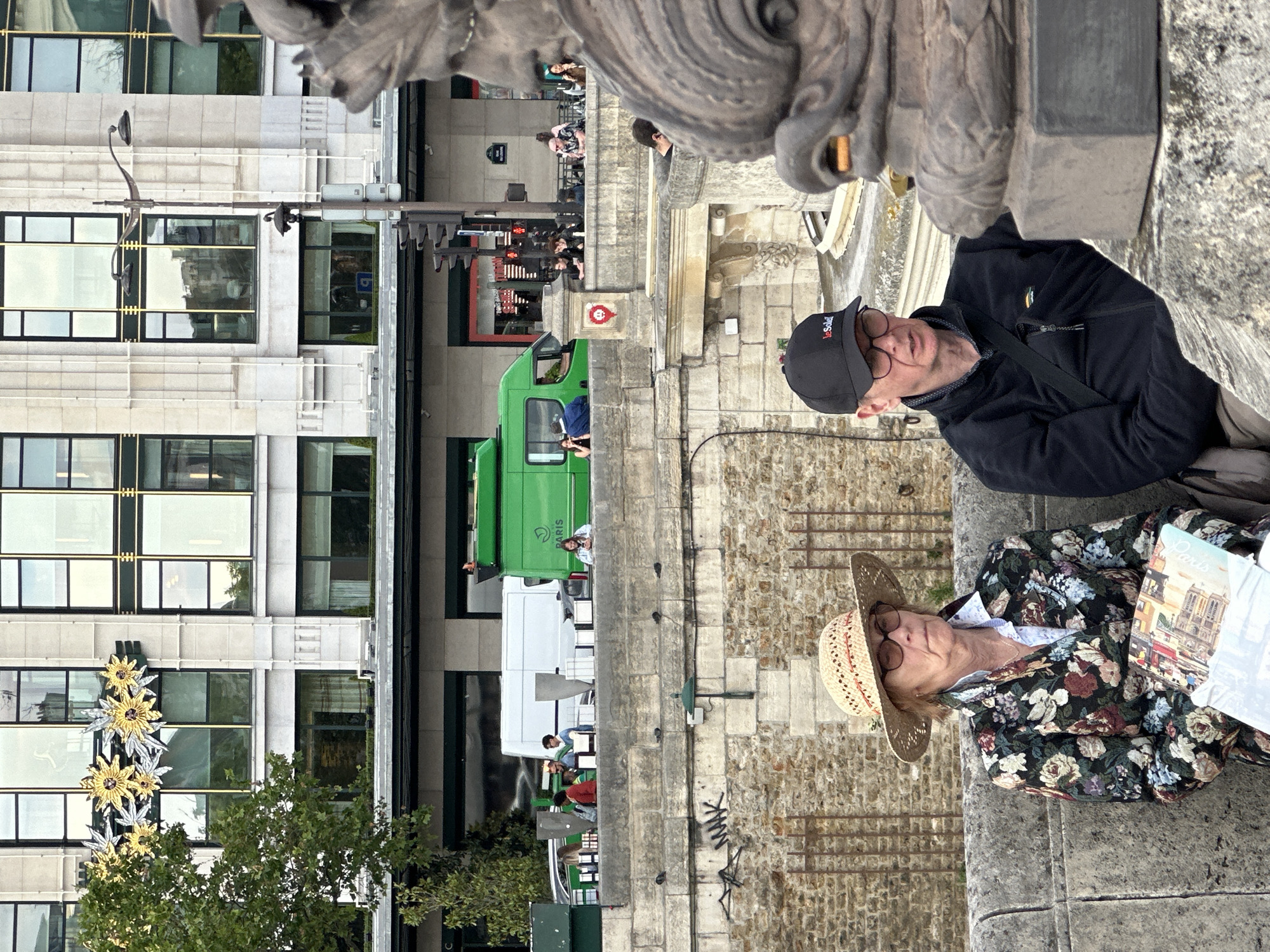 Two people are seated on a stone wall or ledge in Paris, France. One is wearing a floral shirt and straw hat, holding a brochure or map with an image of Notre Dame Cathedral. The other is wearing a black jacket and baseball cap. A green truck is parked on the street below, with pedestrians visible in the background. The building behind the street is beige with black-framed windows and decorative floral ornamentation. The angle of the photograph suggests it was taken on the Pont Neuf bridge.