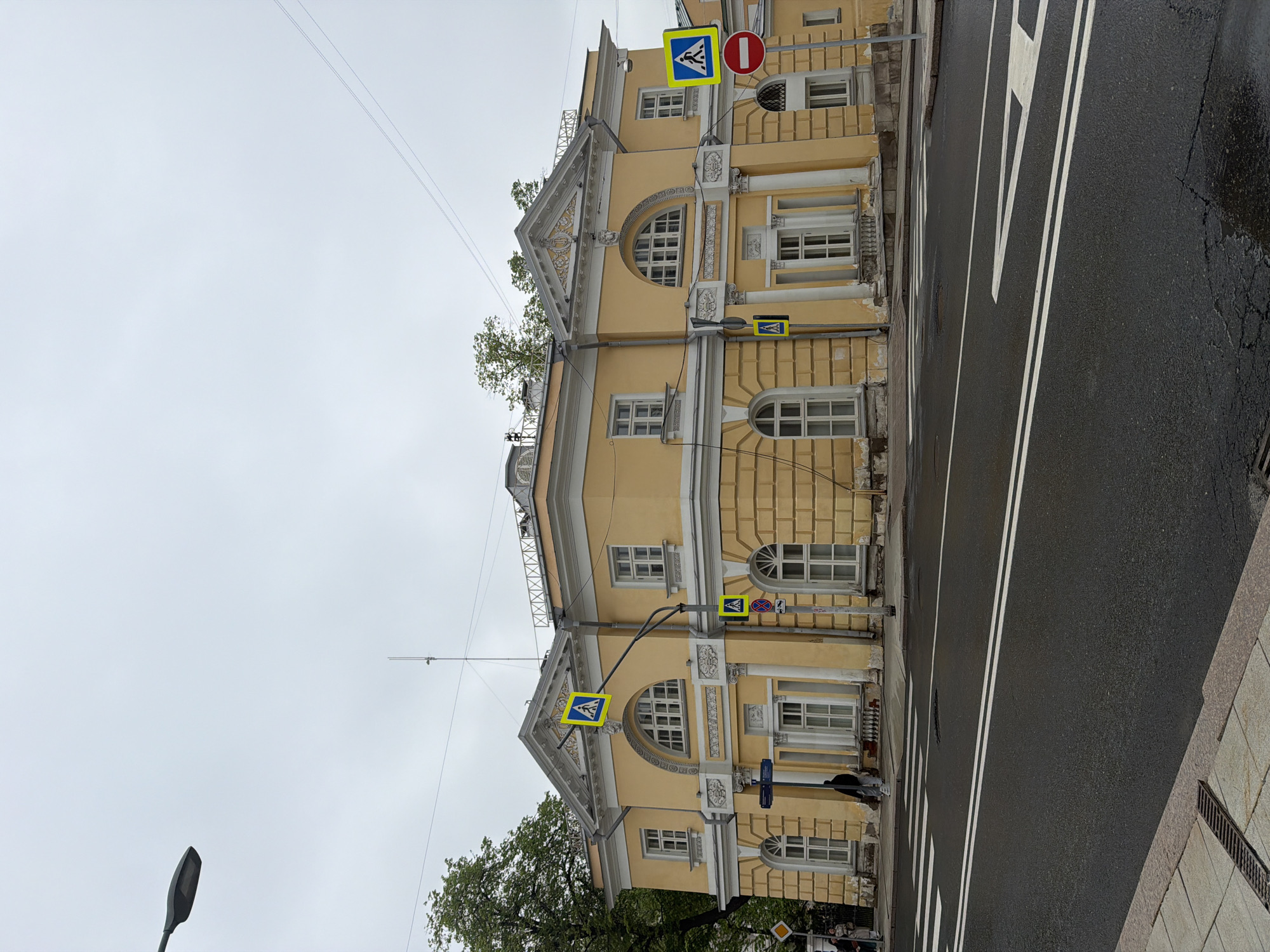 The image shows a three-story yellow building with ornate architectural details. On each level, there are arched windows and rectangular windows. The building faces a street with white lane markings. There are pedestrian crossing signs attached to the building at each level. A street lamp is visible on the left side of the image. The sky is overcast. There is a person walking along the building.