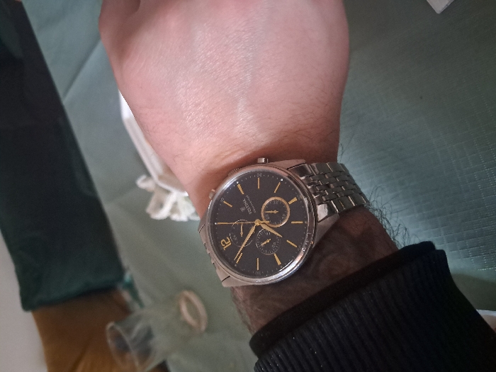 A close-up photograph, taken from a first-person perspective, shows the left forearm and wrist of an individual. The person's skin is light-toned, and they wear a dark, long-sleeved garment with a ribbed cuff on the left arm. A silver-toned "FOSSIL CHRONOGRAPH" wristwatch is prominently displayed on the wrist. The watch features a dark dial, gold-colored hour markers including an Arabic numeral '12', and a metallic linked strap. The arm rests on a light green patterned surface, consistent with a tablecloth. In the soft-focus background, an empty, clear drinking glass and a crumpled white object (likely a napkin) are visible on the table. Further back, parts of dark green and tan/brown upholstered furniture, possibly cushions or a sofa, are discernible. The scene is indoors, in an ambiently lit domestic environment. The location is Unknown, Unknown.