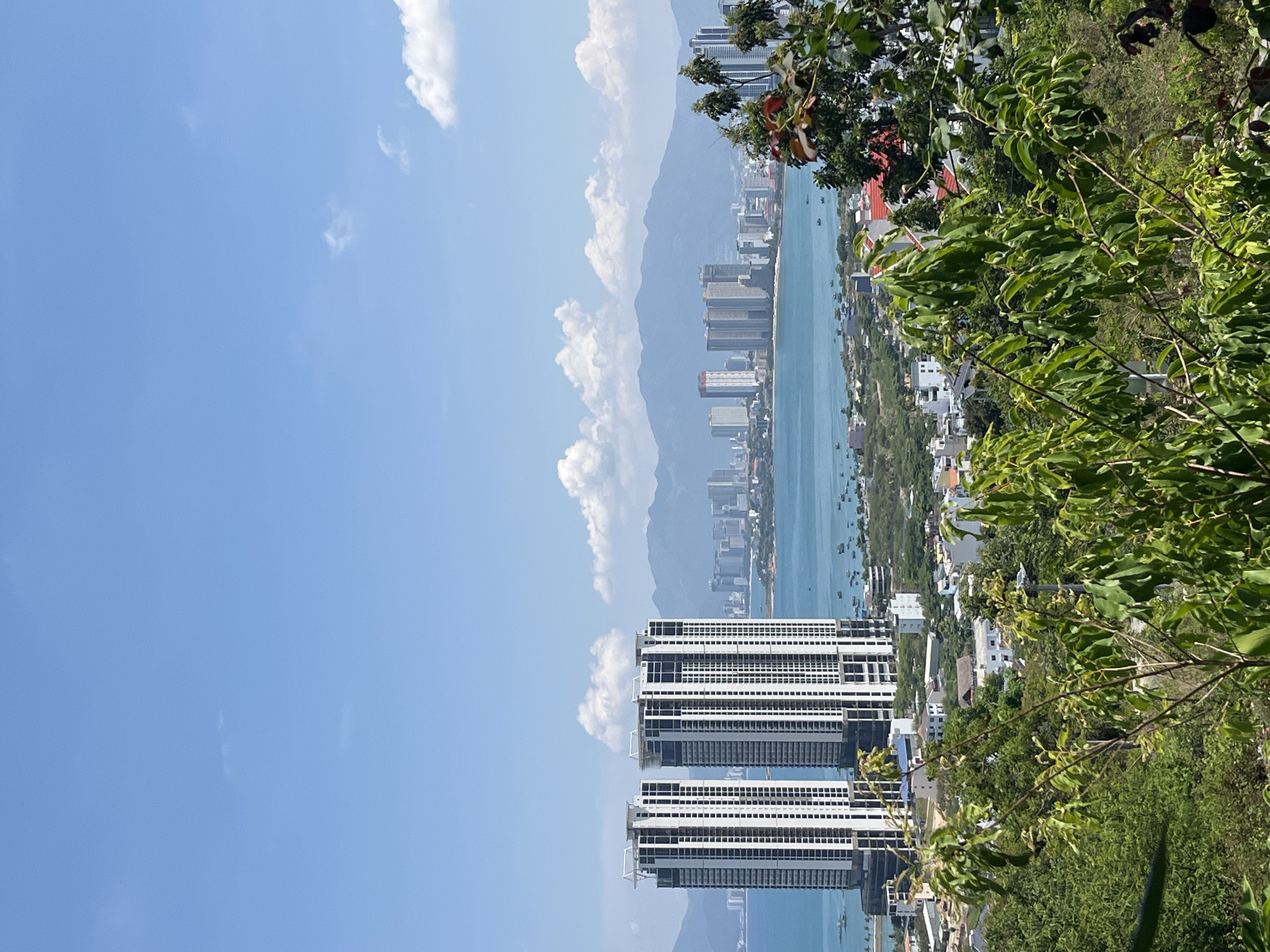 The image is a vertical, elevated view of Bac Nha Trang Ward, Vietnam, shot from an angle looking downwards. The foreground is dominated by dense green vegetation, including trees with vibrant green leaves. A cluster of tall, unfinished buildings with white structures are prominent in the midground, their skeletons partially visible against the clear blue sky and distant landscape. Beyond the buildings, a body of water stretches into the distance, its surface reflecting sunlight. The background features a cityscape with numerous buildings, partially obscured by atmospheric haze and distant mountains. A few clouds are scattered in the upper left portion of the sky.