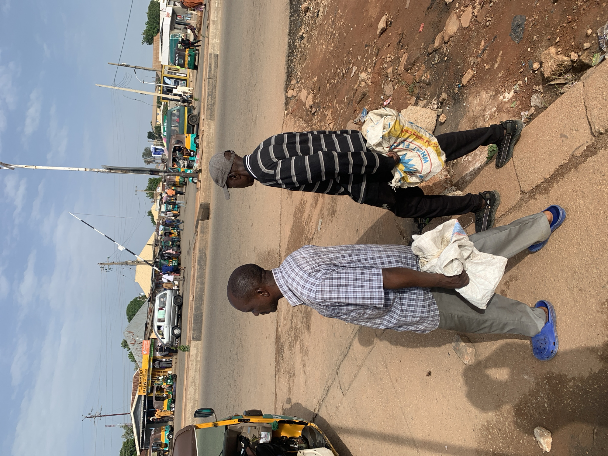 The image shows two men walking on a dirt road in Gura Top, Nigeria, near a busy intersection with several vehicles and buildings. The man on the left, wearing a blue and white checkered shirt and grey pants, is carrying a white bag in his right hand and blue Crocs on his feet. He is walking from left to right. The man on the right, wearing a black and grey striped long-sleeved shirt, black pants, and a grey baseball cap, is carrying a yellow and white bag in his left hand and black sneakers on his feet. He is also walking from left to right. In the background, there are various vehicles including a yellow and green tuk-tuk, a silver van, and a green bus. Several buildings line the road, some with awnings. The sky is partly cloudy.