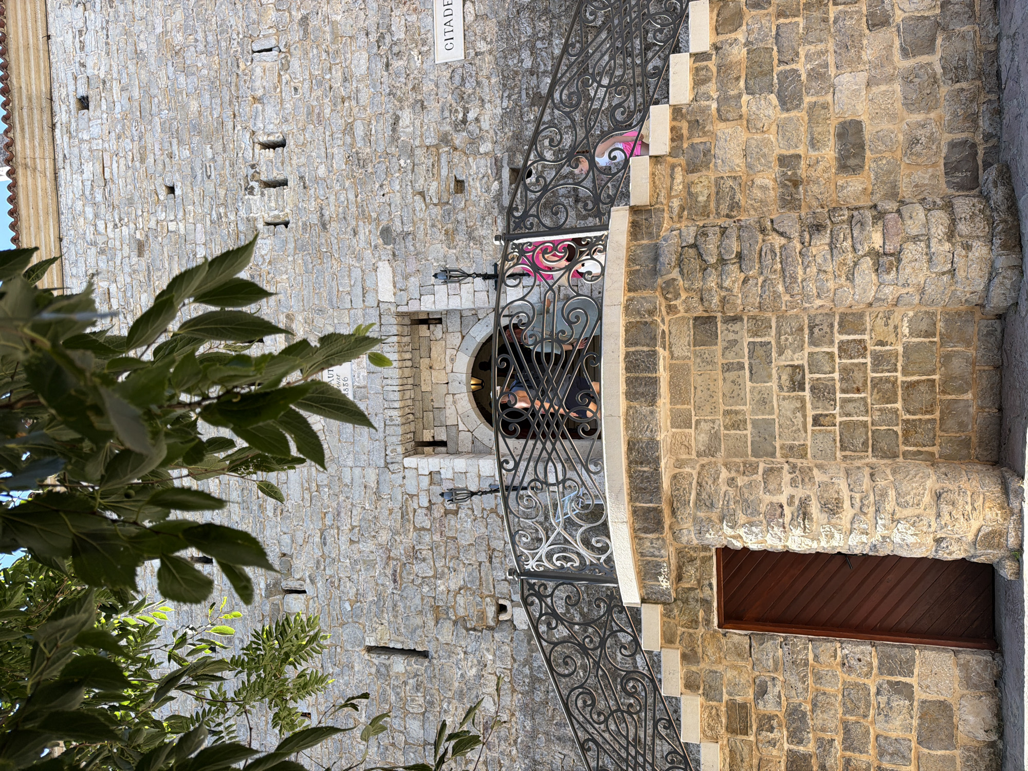The image shows a stone staircase leading up to a fortress, likely part of the Citadel in Budva, Montenegro. Lush green foliage hangs from the top left of the frame, partially obscuring the blue sky. The fortress wall is constructed of rough-hewn stone blocks. At the top of the stairs, an ornate black metal railing lines the walkway. Two ornate black lamps are mounted on the wall near the entrance. Several people are visible on the walkway and stairs. A young girl in a pink skirt and white top is descending the stairs on the right. A woman wearing a dark top and light pants is standing on the walkway, looking into the fortress. Another person is partially visible through the doorway to the left. A wooden door is set into the lower part of the fortress wall on the left side of the image. A sign reading "CITADE" is mounted on the wall to the right of the walkway. The overall impression is one of historical architecture and a sunny day in an urban environment.