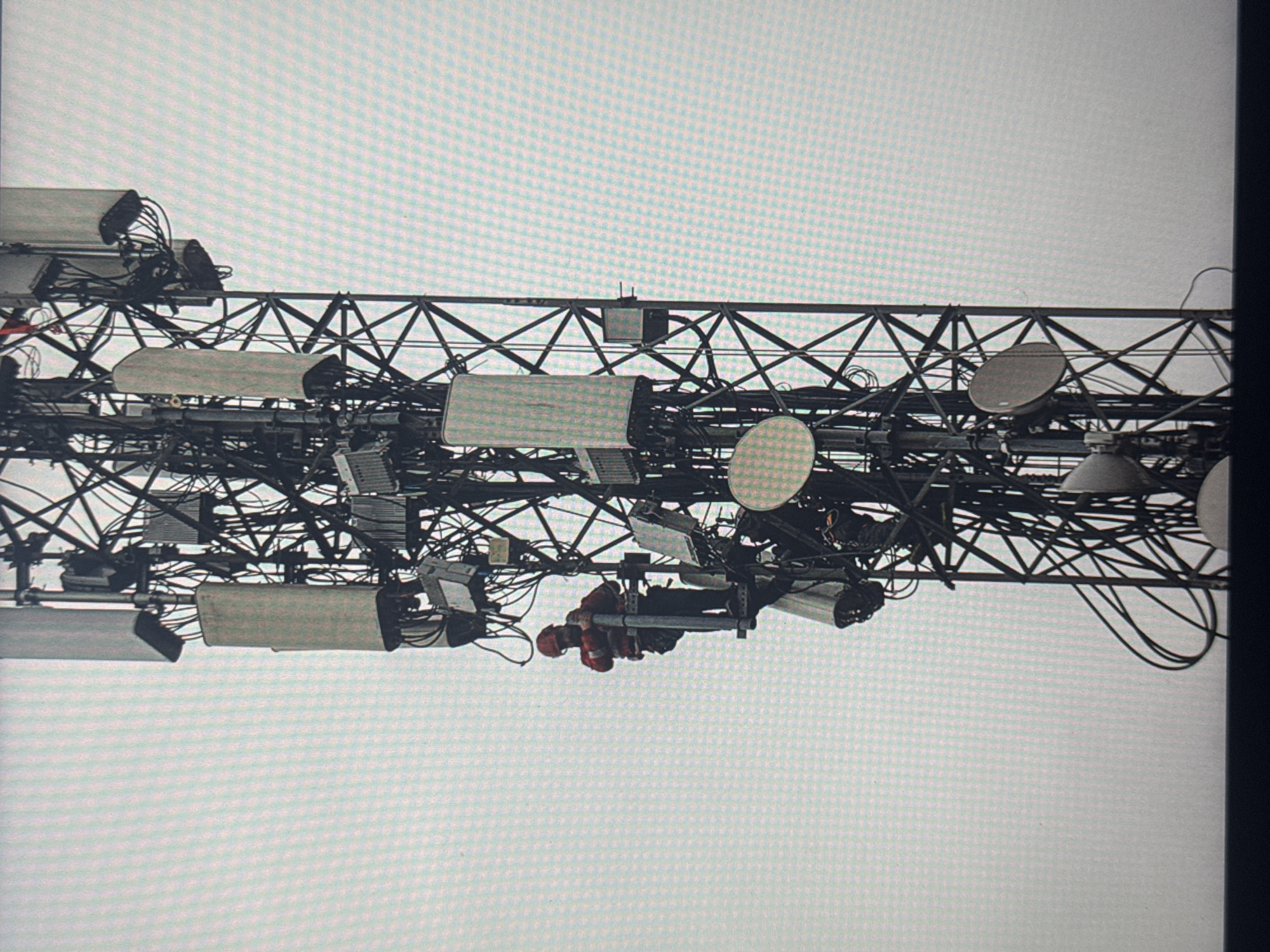 This media file shows a close-up view of a cellular tower, heavily laden with various telecommunications equipment. The structure is a complex latticework of metal beams, with numerous antennas, dishes, and cables crisscrossing in a dense arrangement. Several rectangular devices, likely signal amplifiers or transceivers, are mounted on the tower. Circular satellite dishes are also visible, reflecting the overcast sky.
In the center of the frame, three individuals wearing red safety vests and hard hats are working on the tower. They appear to be engaged in maintenance or installation activities, suspended by harnesses. Their presence suggests that the tower is an active site requiring human intervention.
The lighting is diffused, indicating an overcast or foggy day, typical of many locations. The sky is a uniform light gray. No specific location cues are discernible in the immediate foreground beyond the tower itself. The image is a snapshot of industrial infrastructure and the human labor involved in maintaining it.