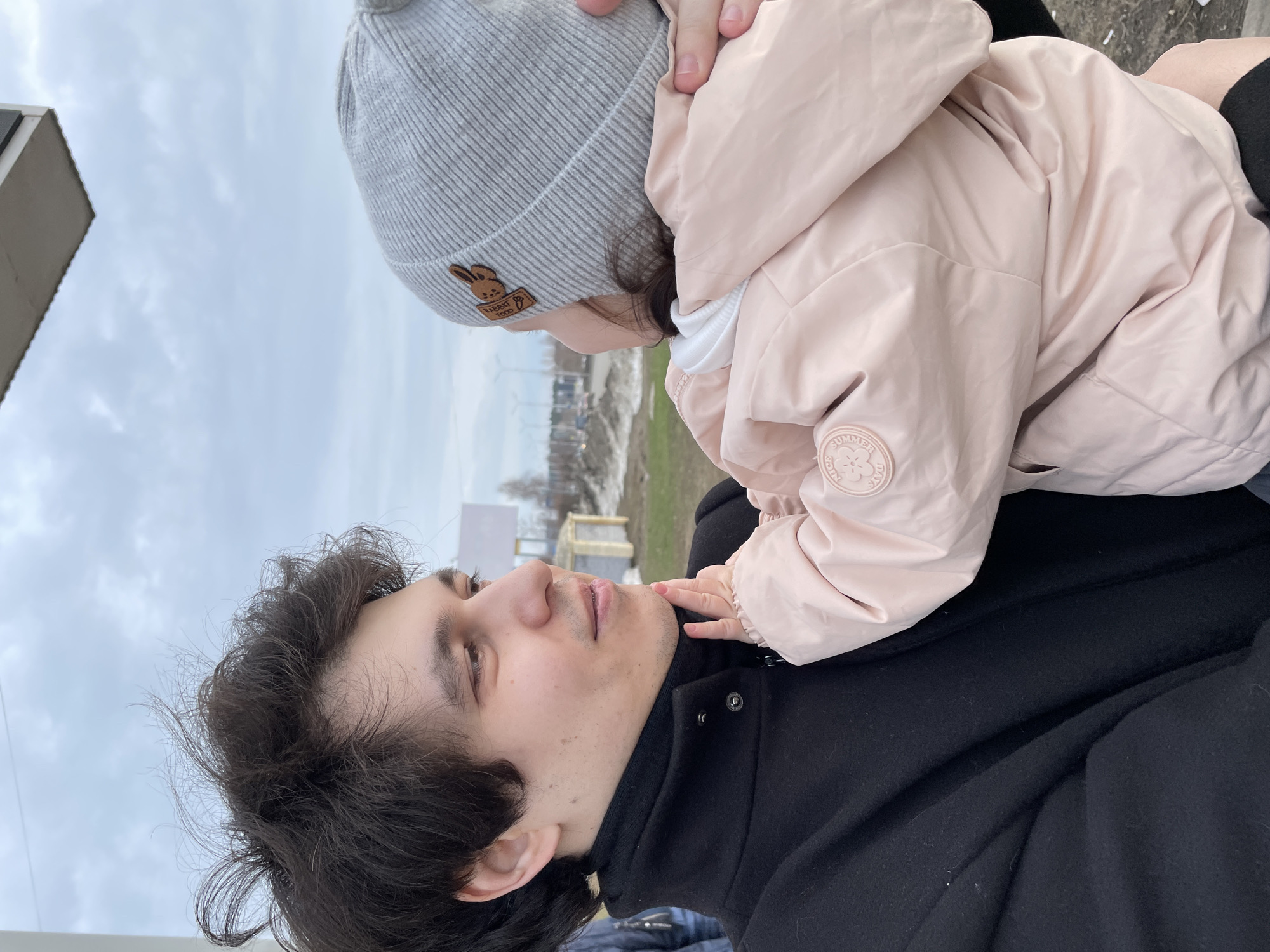 A man and a child are interacting outside. The man is lying on his back, wearing a black coat and looking up at the child. The child, wearing a light pink jacket and a gray knit hat with a bunny patch, is leaning over the man. The child's hand is near the man's face. In the background, there is a cloudy sky, snow on the ground, and some buildings. The location appears to be Izhevsk, Russia, based on visual characteristics.