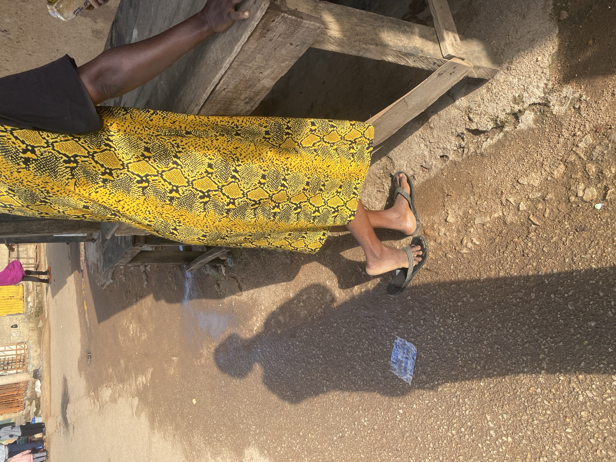 The image captures a street scene in Jos, Nigeria, with a focus on a woman standing near a wooden stall. The woman is wearing a yellow dress with a black and yellow snakeskin pattern and a black t-shirt. She is barefoot, wearing dark-colored flip-flops on her left foot. Her right arm rests on the wooden surface of the stall. A prominent shadow of the woman is cast on the dusty ground to her left, indicating a sunny day. In the foreground, a crumpled piece of blue plastic is visible on the ground within the shadow. The background shows a street with uneven pavement, some puddles of water, and a few other people and structures suggesting a market or commercial area. The overall impression is a snapshot of daily life in an urban environment.