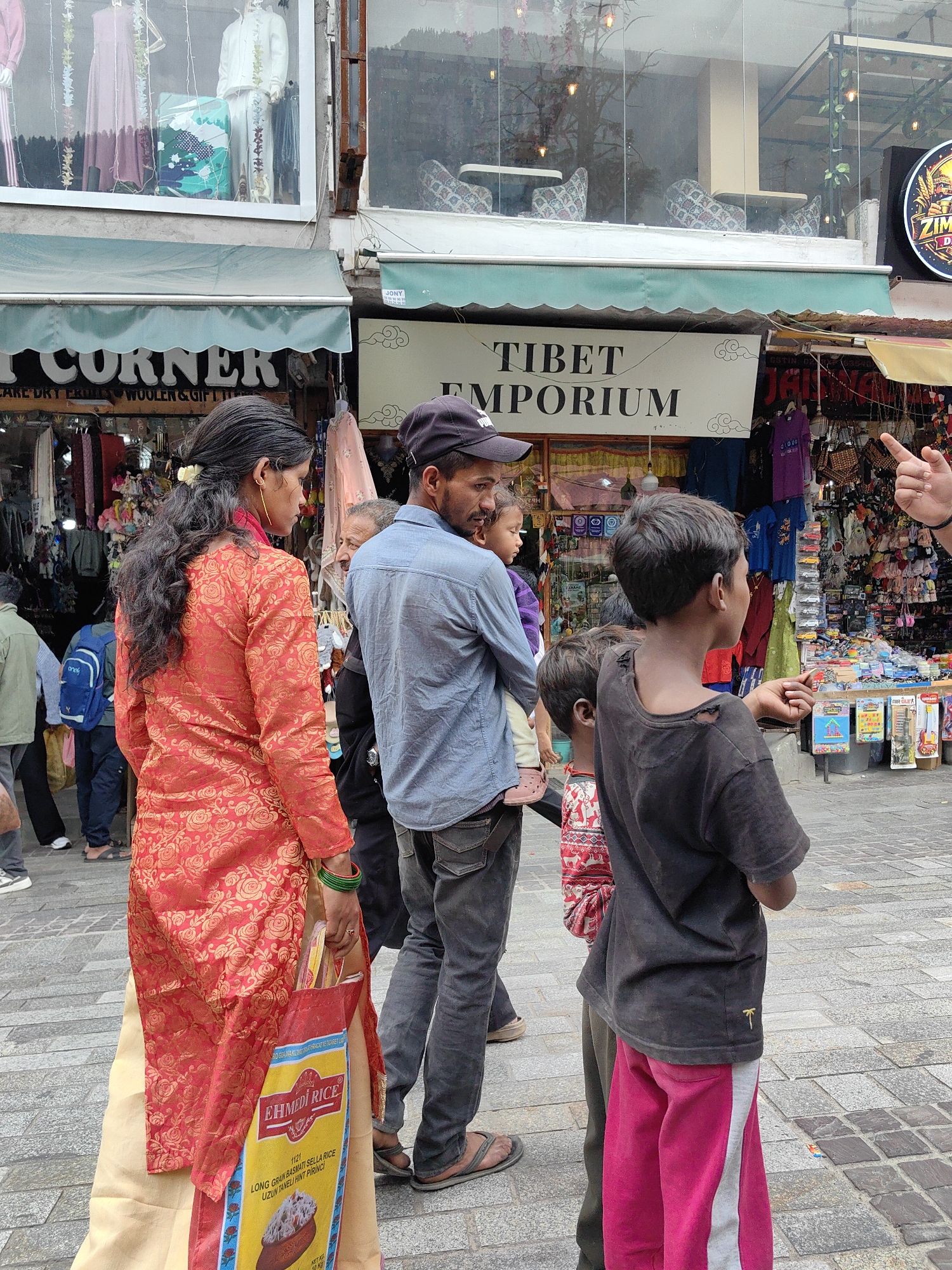 The image captures a street scene in Manali, India. In the foreground, a woman wearing a red and gold patterned kurti and a cream-colored skirt walks while carrying a bag of "EHMEdI RICE". To her right, a man in a blue shirt and dark pants is holding a young child. Beside them, two young boys stand; one in a black t-shirt and pink pants, and the other wearing a red and white patterned shirt. In the background, shops line the street, with signs indicating "CORNER" and "TIBET EMPORIUM". One shop displays mannequins in traditional clothing. The street is paved with stone, and the overall atmosphere suggests a bustling market area.