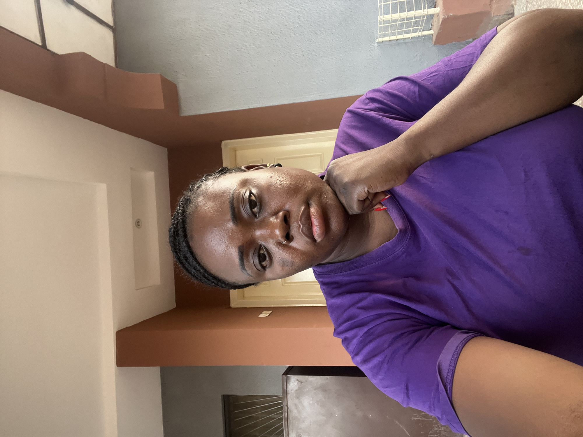 A young adult, with dark skin and braided hair, is depicted in a close-up selfie, filling the left and center portions of the frame. She wears a vibrant purple t-shirt and has a nose piercing. Her left hand is casually resting on her chest or shoulder, with fingers slightly clenched. She gazes directly at the camera with a neutral to slightly contemplative expression, her lips gently parted.
The background reveals an interior or semi-enclosed architectural space, possibly a hallway or covered porch. Distinct architectural elements include a light beige door and door frame, a prominent brown or terracotta-colored structural beam or overhang, and walls painted in light blue-grey and off-white shades. A white grid-like structure, possibly a shelf or ventilation grate, is visible in the upper right. Another metal grate and a darker surface are partially visible in the lower left background. The lighting suggests it is daytime. No visible text is present in the image. The scene is located in Jos, Nigeria.