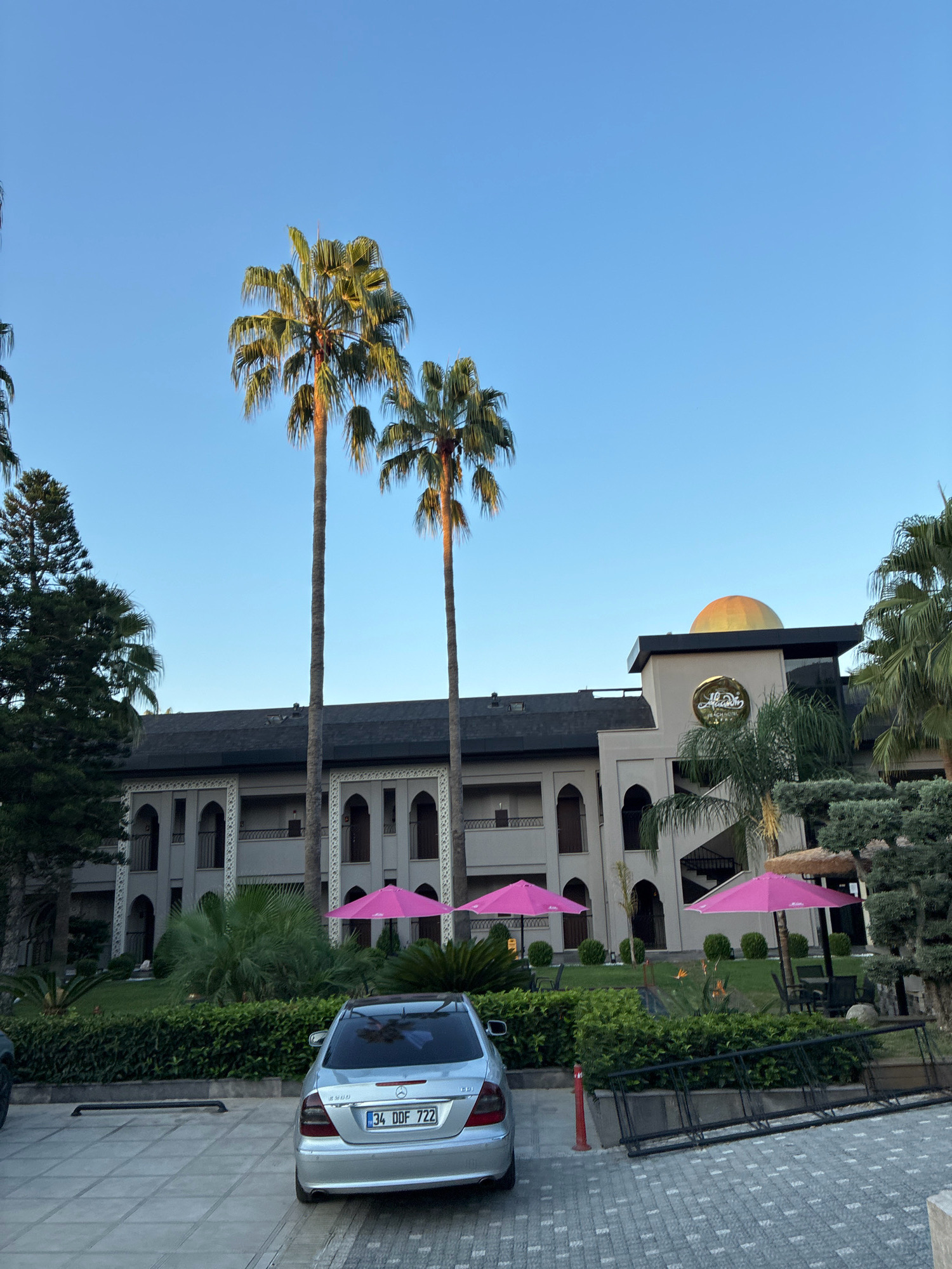 The image depicts the front facade of the Aladdin Beach Hotel in Alanya, Turkey, under a clear blue sky. The hotel exhibits a distinctive architectural style, featuring a central golden dome atop a beige structure, with a prominent sign displaying "Aladdin Beach Hotel." The building is characterized by its arched windows and balconies, set against a dark roof. In front of the hotel, several vibrant pink umbrellas are positioned over seating areas.
Two tall palm trees stand prominently in the foreground, flanking the hotel. To the left, a dense conifer tree adds greenery to the scene. A silver Mercedes-Benz sedan is parked on a paved driveway in front of the hotel. The surrounding area is landscaped with manicured bushes and lush green grass. In the lower right corner, a metal ladder lies on the ground. The overall impression is that of a well-maintained resort, captured during daylight.