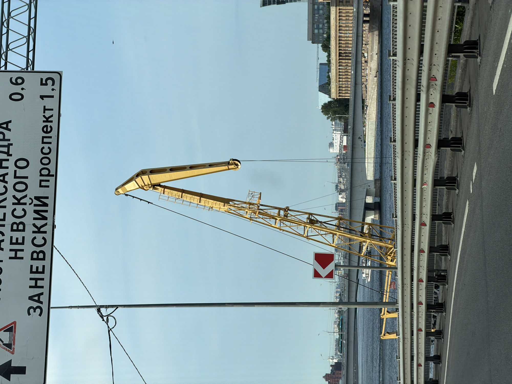 The image shows a street view in Saint Petersburg, Russia, focusing on a road with a guardrail, a large yellow crane, a body of water with a bridge and boats, and buildings in the background. A traffic sign in Russian indicates directions to "NEVSKY PROSPEKT" at 1.5 km and "ALEKSANDRA NEVSKOGO" at 0.6 km. The crane is positioned near the water, suggesting a port or industrial area. The sky is clear and blue. There are no people visible in the foreground. The overall impression is of an urban environment with infrastructure related to transport and industry.