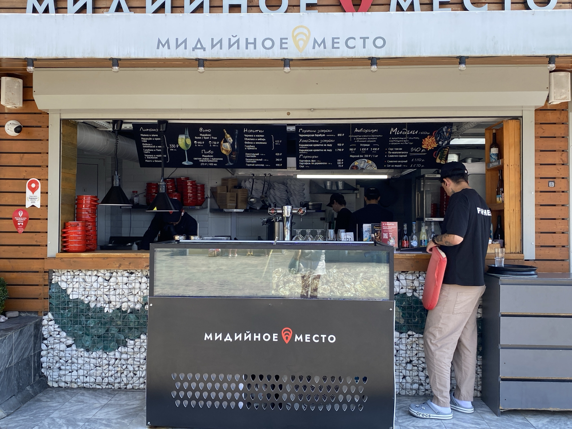 The image shows a street food vendor called "Мидийное место" (Midiynoye Mesto), which translates to "Mussel Place," located in Sochi, Russia. The vendor is a small kiosk with a counter and a menu displayed above. A person wearing a black t-shirt and light-colored pants stands behind the counter, seemingly serving a customer. The kiosk is decorated with stones and has a prominent logo with the name of the establishment. The surrounding area appears to be an outdoor public space, possibly a promenade or a street with other businesses. The weather is sunny, suggesting a pleasant day.