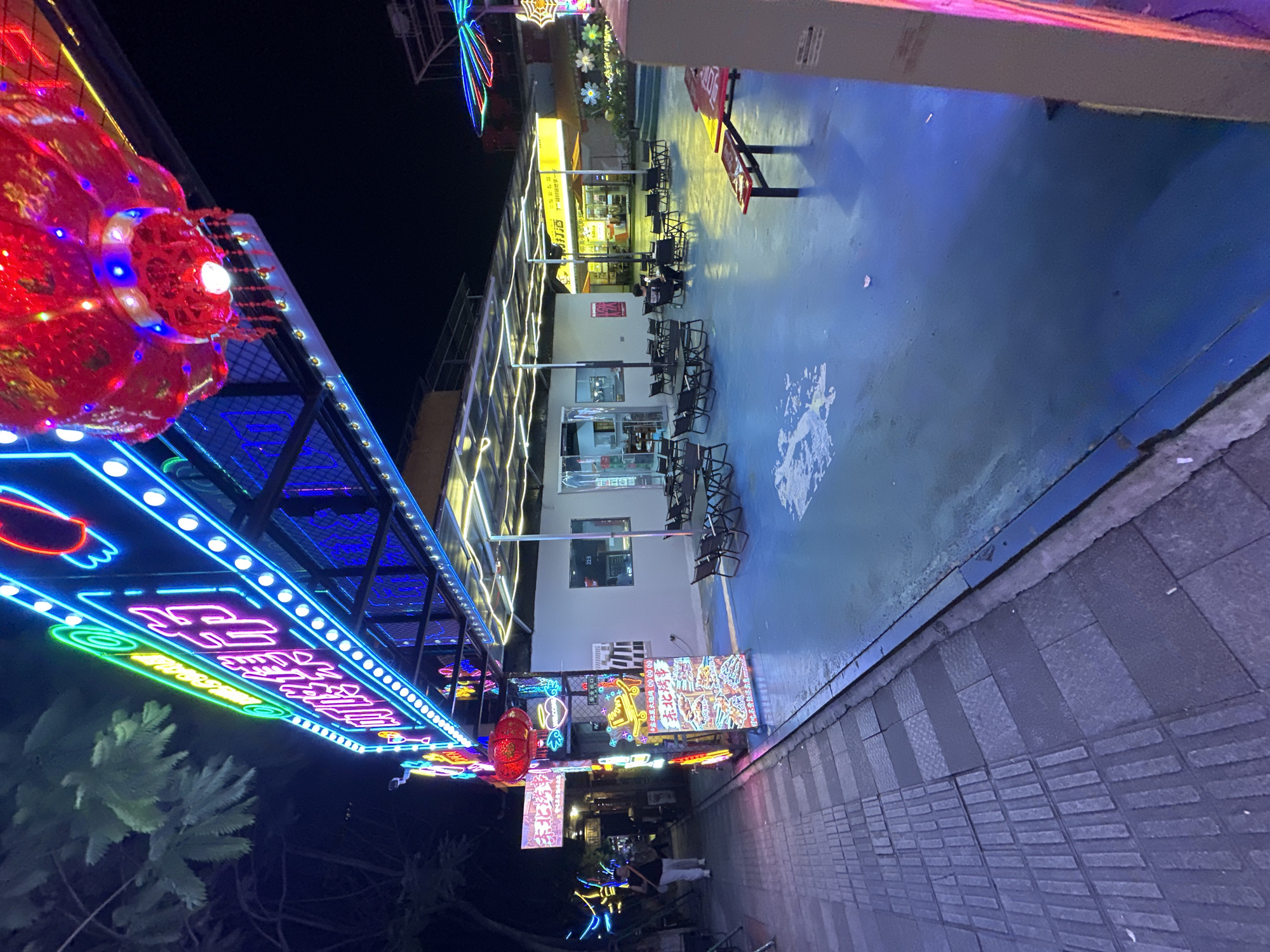 This is a night-time, eye-level, outdoor shot taken on a paved area in the city of Chongqing High-tech Zone, China. The image depicts a vibrant street scene, likely an entertainment or dining area. Dominating the upper portion of the frame is a brightly lit neon sign that reads "東北烤串" (Dongbei Kaoxuan - Northeast Skewers) in Chinese characters, illuminated with pink and blue neon lights, surrounded by white circular bulbs. To the right of the sign, a large, red, decorative lantern hangs, also lit, with intricate gold patterns.
Below the neon sign, a covered area with a transparent roof stretches across the scene. This area has multiple tables and chairs set up, indicating it's a seating area for patrons. The ground within this area is a striking blue, with some white patterns or reflections on its surface. A few people are visible: one person is walking on the left side of the image on a gray tiled sidewalk, wearing light-colored pants and a dark top. Another person is seated at a table within the covered area.
Further into the background, more buildings are visible, some with yellow illuminated signs. One sign appears to read "好店" (Hao Dian - Good Store). Decorative lights, including what looks like a colorful, starburst-shaped light fixture, add to the festive atmosphere. The overall environment suggests a lively commercial street or food court illuminated by artificial lights. The image is sharp and well-exposed, capturing the details of the neon signs and the surrounding structures.