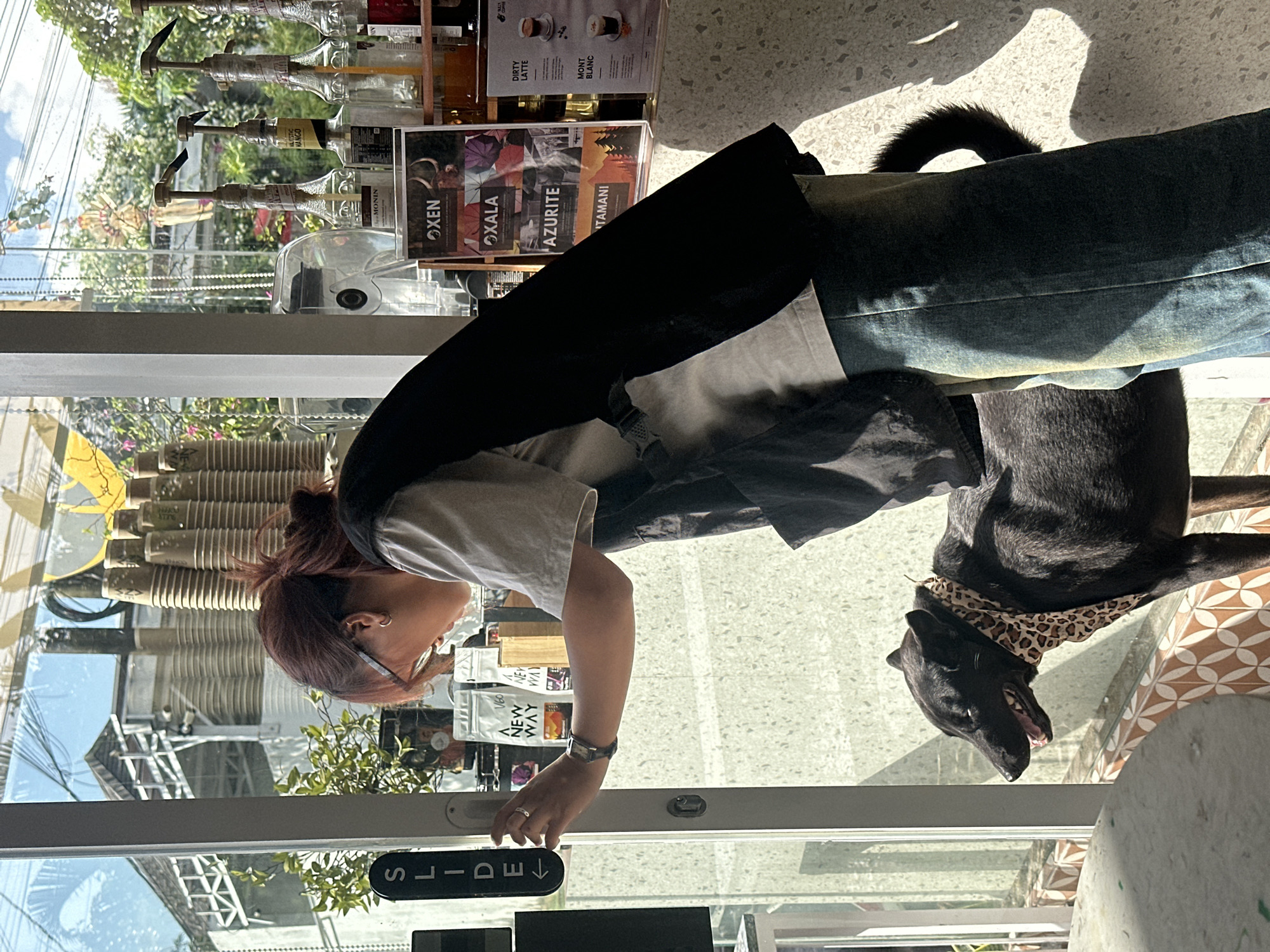 A person with reddish-brown hair pulled back in a bun is leaning forward, looking down and to the left through a glass door, with their left hand on the door handle, which is next to a black sign with white text that says "SLIDE" and an arrow pointing left. They are wearing glasses, a light-colored t-shirt, a dark vest, and blue jeans. A black dog with a leopard-print bandana around its neck stands to the person's left, looking to the left with its mouth slightly open. Sunlight streams in from the right, casting shadows. Behind the person and to the right, on a counter, are several glass bottles with pumps, stacks of paper cups, and various signs and packaging. The background visible through the glass door shows a building with white architectural elements and greenery. The scene appears to be indoors at a cafe or shop in North Kuta, Indonesia, given the lighting and architectural style visible through the glass.