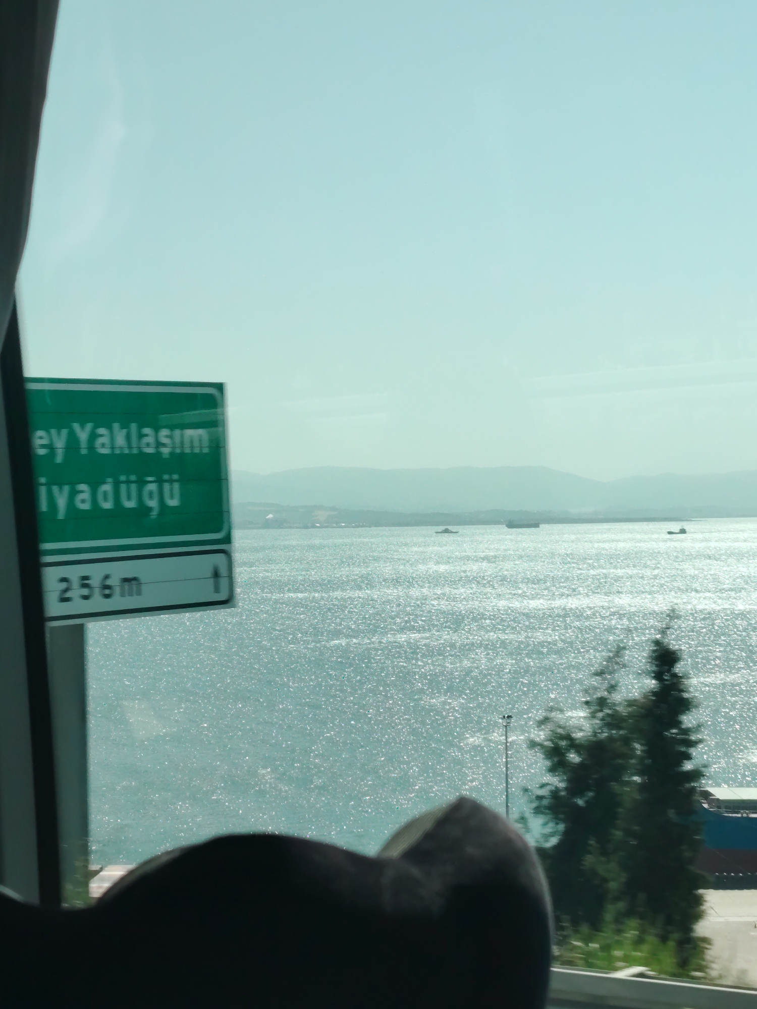 The image captures a bright, hazy daytime scene looking out over a wide body of water, likely the Gulf of Izmit near Dilovası, Turkey, from inside a vehicle. The viewpoint is from a passenger seat, with a dark, blurred object resembling a headrest in the foreground.
On the left, a prominent green road sign with white text is partially visible. The readable text includes "ey Yaklaşım" and "iyadüğu" (likely part of a place name or instruction), followed by "256m" and an upward-pointing arrow, indicating a distance to a destination or turn.
The main subject is the sparkling, turquoise-blue water, which is extensively covered in bright, shimmering reflections of sunlight, suggesting a clear but possibly hazy sky. In the far distance, a soft, hazy coastline with mountains or hills stretches across the horizon. Several vessels, including what appears to be a large cargo ship and smaller boats, are visible on the water.
To the right, some dark green trees or foliage are seen, partially obscuring the view of the water and what could be a part of a port or industrial area, with a large blue structure visible. The sky above is a light, hazy blue. The scene depicts travel, possibly by bus or car, along a road adjacent to the sea or a large bay.