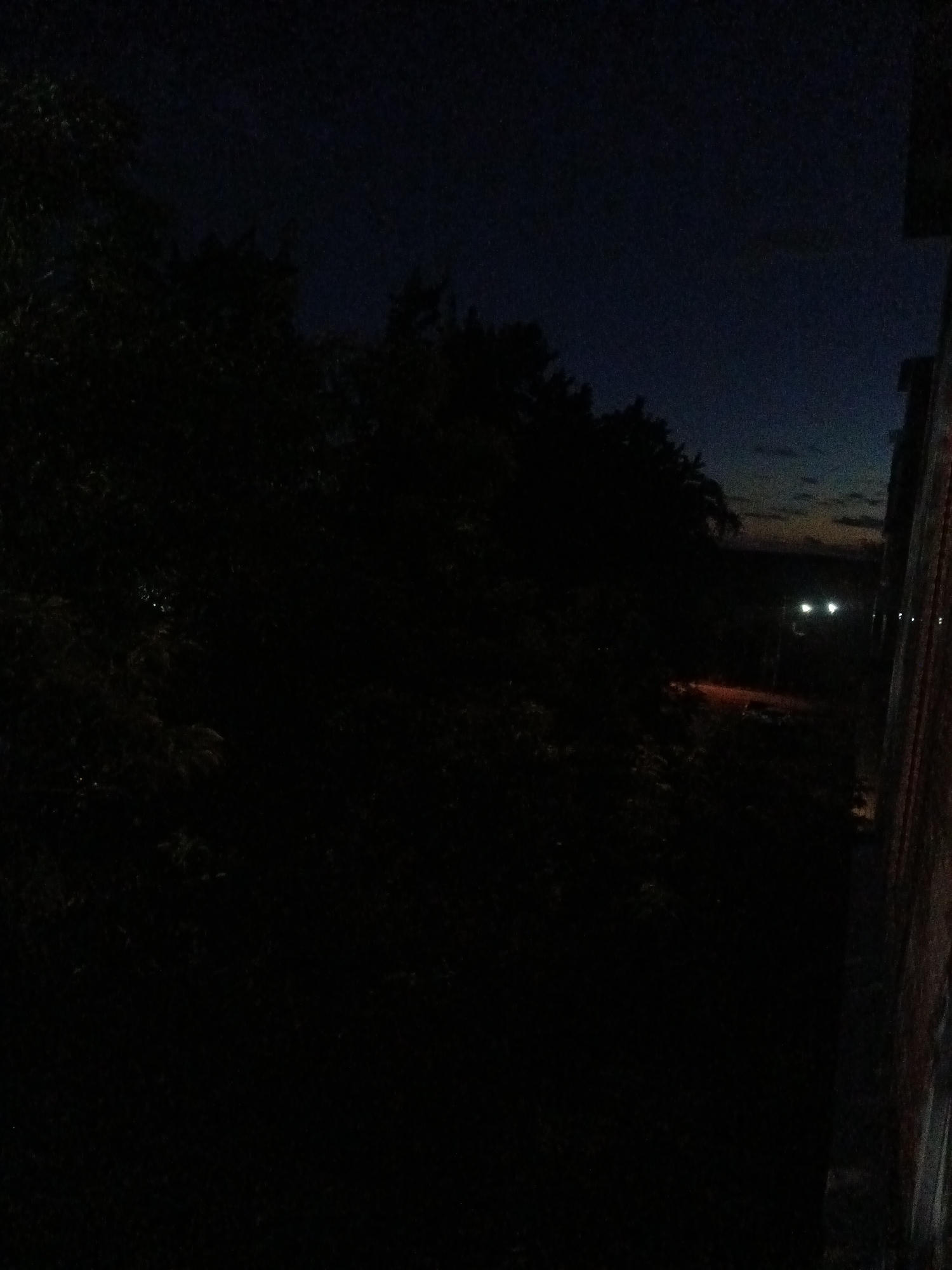 The image depicts a dark, possibly twilight or night scene with a dimly lit outdoor environment. In the foreground and left side of the frame, dense foliage of trees is visible, appearing as dark silhouettes against a darker sky. The sky itself is a deep navy blue, with a hint of lighter colors near the horizon on the right, suggesting the last vestiges of daylight or distant city lights.
Further into the scene, on the right side of the image, the edge of a building is visible. It has a rough, textured surface, possibly brick or concrete, with vertical lines that suggest construction or weathering. Adjacent to this building, a road or path can be inferred, with a warm, orange-red glow emanating from it, possibly from streetlights or a vehicle. Two bright, white headlights of a vehicle are also visible in the distance, moving away from the viewer. A few other faint, distant lights are also present.
The overall impression is one of a quiet, possibly suburban or semi-rural area on the outskirts of a city, illuminated by the remaining ambient light of the sky and artificial light sources. The location is specified as Kuznetsk, Russia. There are no people clearly visible in the frame.