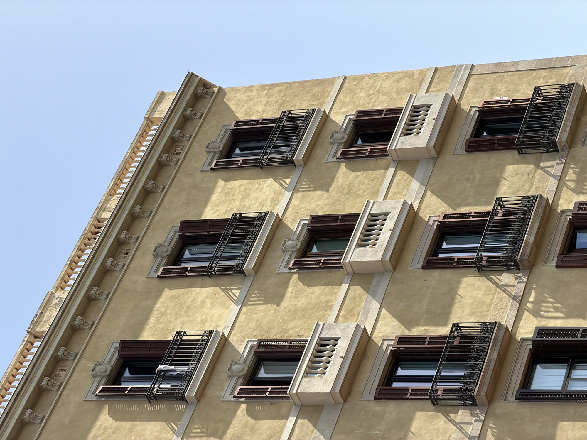 This is a close-up, eye-level, full-shot image of the facade of a building in Barcelona, Spain. The building is constructed with yellow stucco and features ornate architectural details such as decorative moldings and a balustrade along the roofline. There are multiple windows with dark brown wooden shutters, some of which are open, revealing glimpses of the interior. Each window also has a small, wrought-iron balcony. The sunlight casts shadows on the building, indicating a bright, clear day with a light blue sky. There are no people visible in the image.