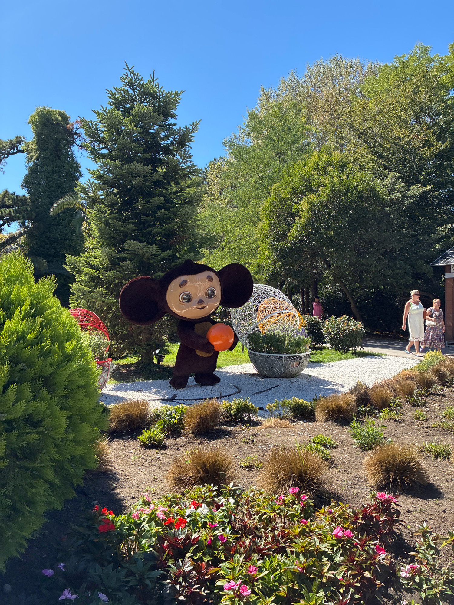 This image captures an outdoor scene in Sochi, Russia, likely within a park or garden. In the foreground, a large, plush Cheburashka character, a popular Russian cartoon character, stands holding an orange ball. To the left, a vibrant green coniferous bush is prominent. The mid-ground features a pathway lined with white pebbles, leading past a decorative wire sculpture containing green foliage. Several people are visible in the background: two women in light-colored dresses walk along the pathway, and a child in a pink shirt stands near some trees. Lush green trees and foliage dominate the background, with a clear blue sky overhead. The overall impression is a sunny day in a well-maintained outdoor recreational area.