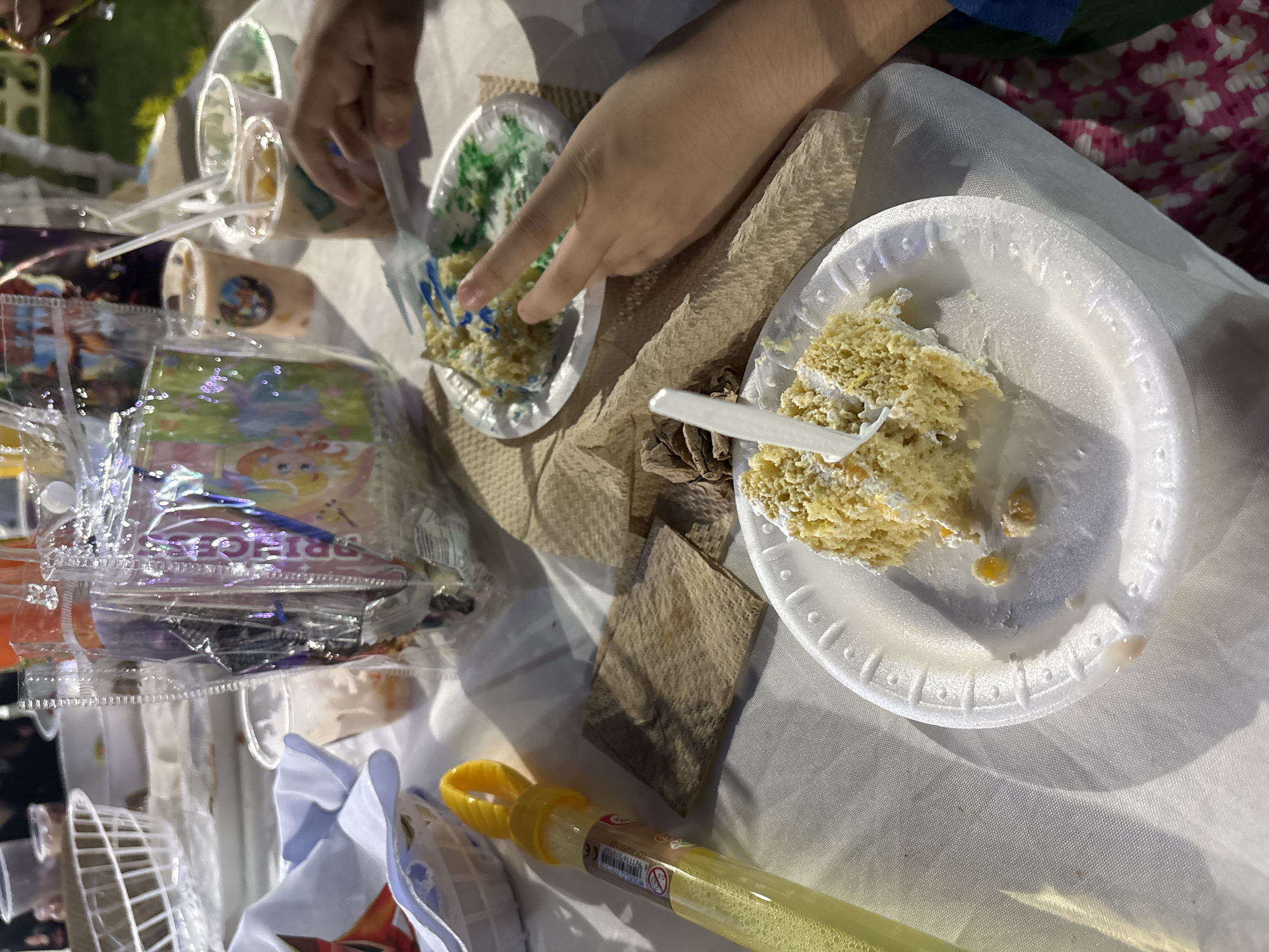 The image depicts a close-up view of a white plastic table covered with a white tablecloth. On the table, there are two plates with slices of cake. The cake on the foreground plate is yellow with white frosting and some yellow pieces, and it is partially eaten. A white plastic fork is stuck into this cake. Another plate with a slice of cake, partially eaten and decorated with green and blue frosting, is visible in the background. A person's hand is reaching towards this second plate, with their index and middle fingers extended. To the left of the foreground plate, there is a yellow bubble wand with a yellow loop and a clear tube filled with yellow liquid, presumably bubble solution. Several paper napkins are scattered on the table, some appearing used. In the background, there are plastic cups with drinks, one with a straw, and a clear plastic bag containing what appears to be party favors with images of cartoon characters. The scene suggests a casual outdoor gathering, possibly a child's birthday party, given the presence of cake, party favors, and bubble wand. The lighting suggests it may be late afternoon or evening, with shadows cast on the tablecloth. The context of Progreso, Mexico, suggests this is likely a beachside or outdoor event in that location.
