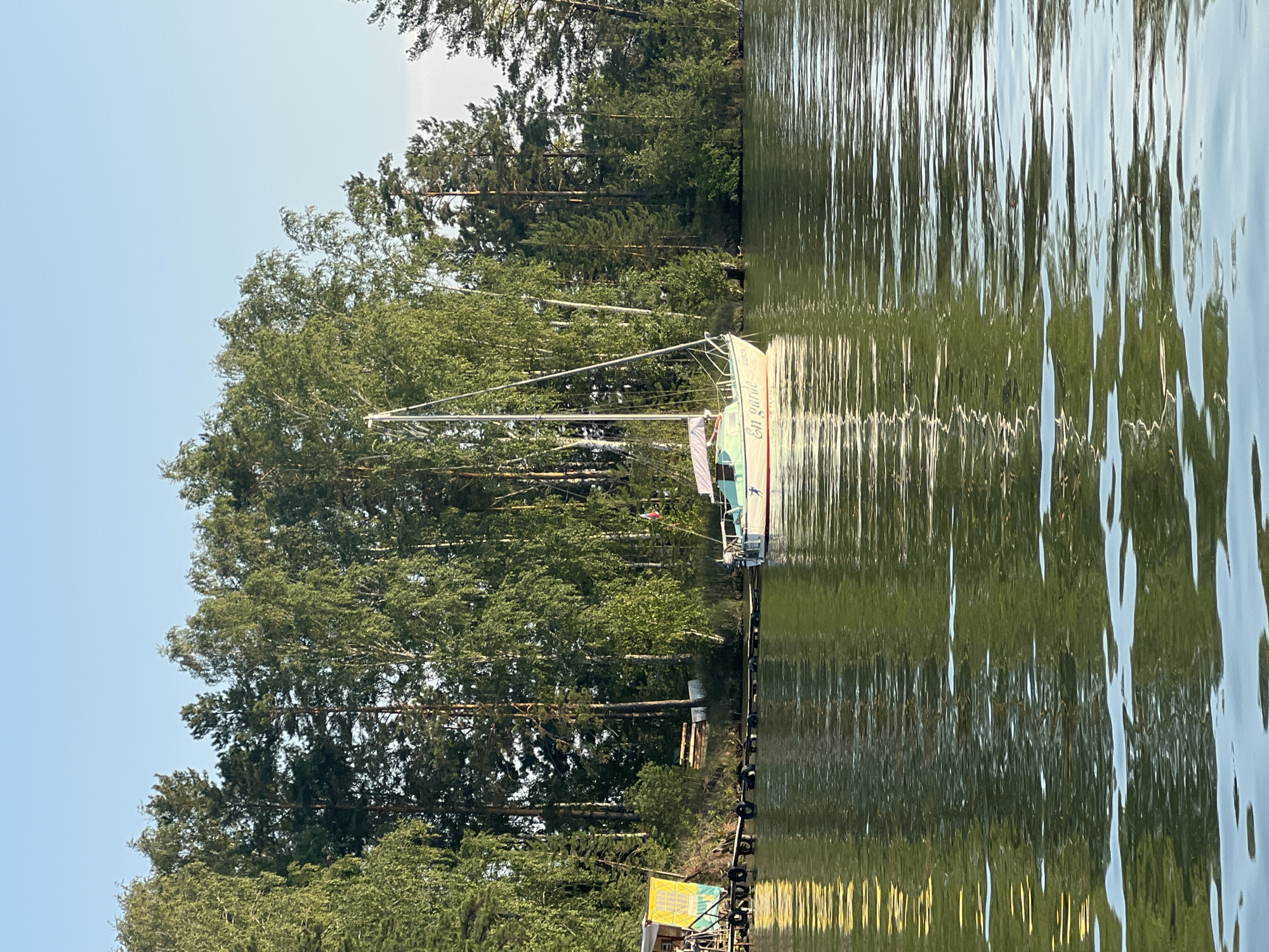 The image shows a serene lake scene with a sailboat docked near a wooden pier on the shore. The sailboat, named "En garde," is white with a teal stripe and a white sail furled. It is positioned in the calm, green water, reflecting the surrounding trees. The shore is lined with lush green trees, primarily birch and pine, under a clear blue sky. To the left of the sailboat, a small, yellow and blue structure with a slanted roof is visible, connected to the pier. The pier itself is made of dark wood and is lined with black tires, likely serving as fenders for docked boats. The water exhibits gentle ripples, catching the sunlight and reflecting the greenery and the sky. The overall impression is one of a peaceful, natural setting. The city of Borovoe, Russia, is known for its picturesque lakes and surrounding forests, making this scene consistent with the natural beauty of the region.