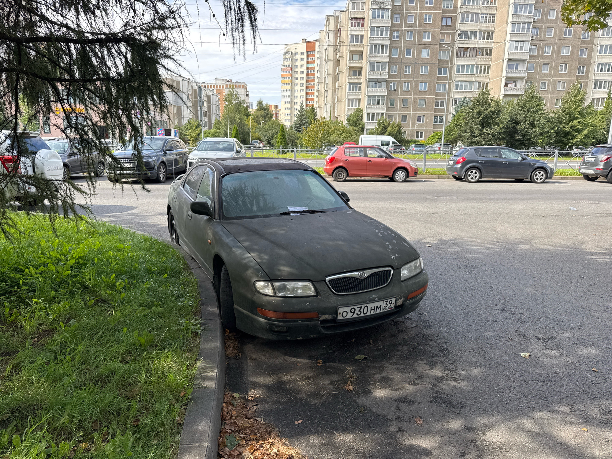 In the city of Kaliningrad, Russia, on a sunny day, a dark green sedan with a license plate reading "O 930 HM 39 RUS" is parked on the side of a street. The car is positioned next to a curb with grass growing on the other side. Behind the sedan, a red hatchback, a dark gray SUV, and other vehicles are visible, parked along the street. In the background, tall apartment buildings stand, with trees and a cloudy sky above. The pavement is asphalt, with some fallen leaves scattered around.