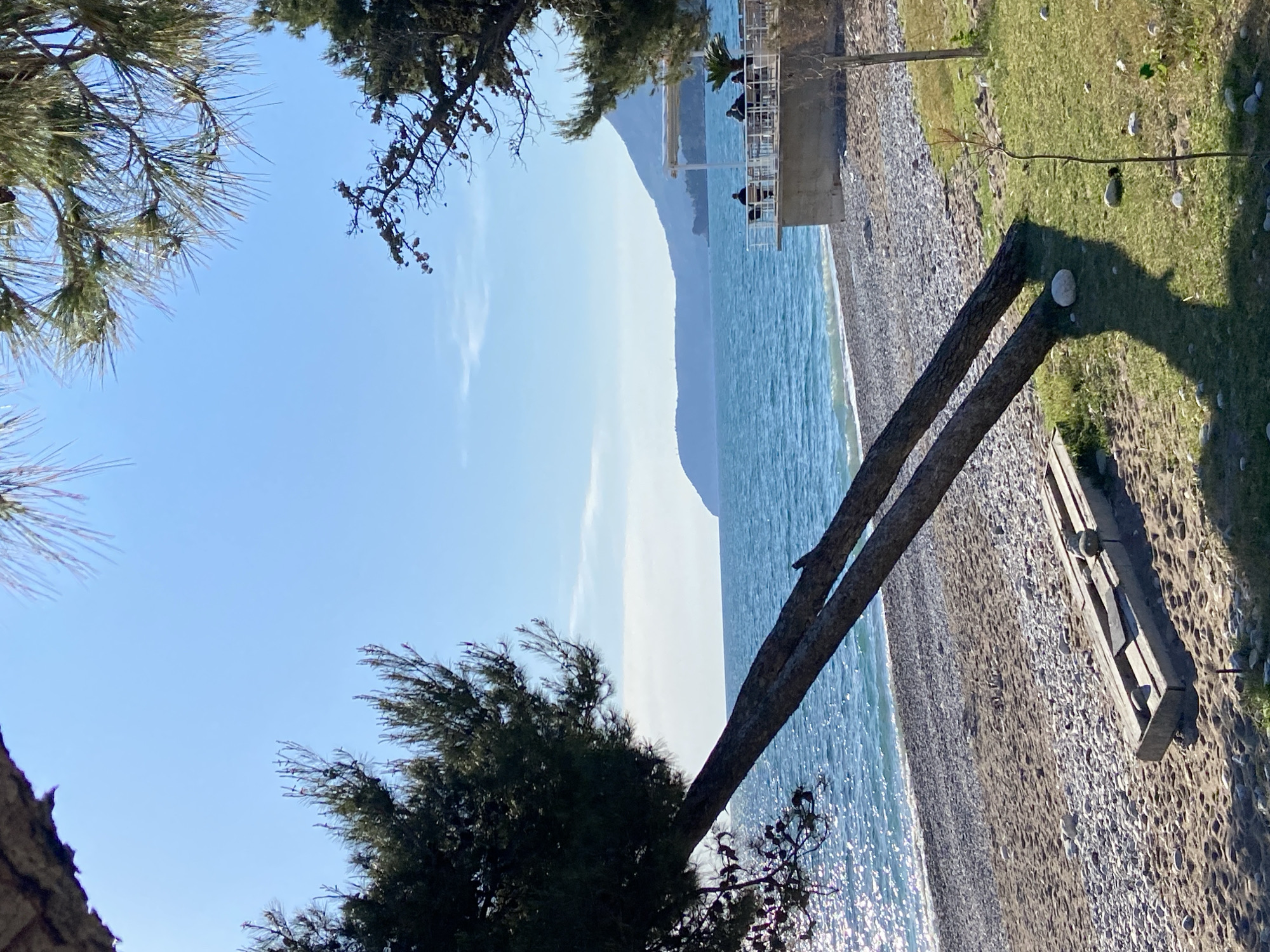 The image shows a beach scene in Bar, Montenegro. The sea occupies the central part of the image, with visible light reflecting on the water's surface. A pebble beach is visible in the lower right, where a wooden beach bench rests. To the right of the bench, grass occupies a thin strip. On the beach's border, a couple of people are sitting on a concrete balcony overlooking the water. Trees frame the image's top and left sides. A distant mountain is visible across the water. The sky is blue with a few faint clouds.