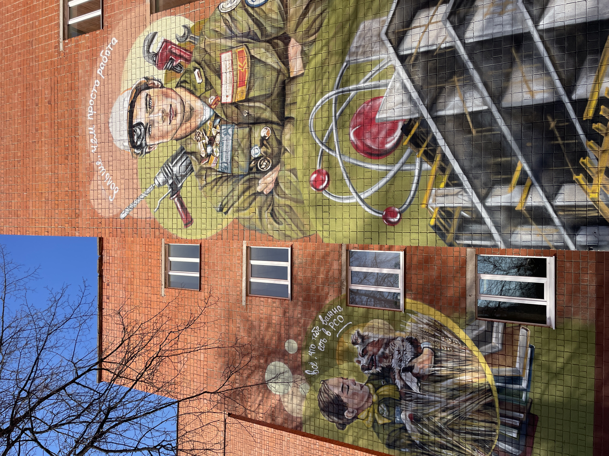 The image shows a mural painted on the side of a brick building. In the top portion of the mural, a figure wearing a white cap and goggles is depicted in an olive-green uniform, adorned with medals and holding a drill. Next to the figure is an atom model. The text, written in white cursive is present. Further down on the mural is a second figure with a raccoon on their shoulders. The text, written in white cursive is present. The mural is on the exterior wall of a building with several windows. The sky is visible in the lower left corner, with bare tree branches in the foreground. The image is oriented sideways. The location is Izhevsk, Russia.