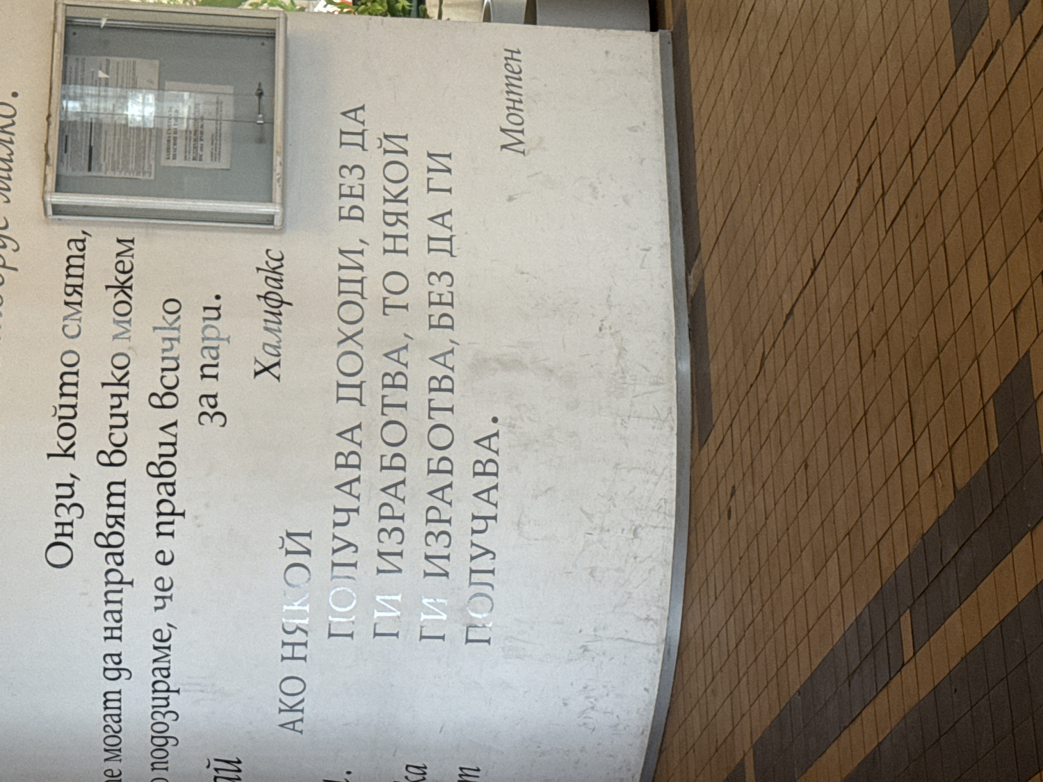 The image, taken at an oblique angle, shows a light-colored wall adorned with two Bulgarian text inscriptions, and a section of a tiled floor. On the left side of the wall, there is a small, glass-fronted notice board, but its contents are obscured by glare.
The text on the left side of the wall reads:
"Онзи, който смята, че могат да направят всичко за пари, подозираме, че е правил всичко за пари.
Халифакс"
This translates to: "The person who thinks that they can do anything for money, we suspect that they have done everything for money. Halifax"
The text on the right side of the wall reads:
"АКО НЯКОЙ ПОЛУЧАВА ДОХОДИ, БЕЗ ДА ГИ ИЗРАБОТВА, ТО НЯКОЙ ГИ ИЗРАБОТВА, БЕЗ ДА ГИ ПОЛУЧАВА.
Монтен"
This translates to: "IF SOMEONE RECEIVES INCOME WITHOUT EARNING IT, THEN SOMEONE EARNS IT WITHOUT RECEIVING IT. Montaigne"
The wall itself appears to be made of a smooth, light-grey material, possibly concrete or stone. Below the wall and to the right, a floor made of square tiles is visible. The tiles are primarily brown, with some darker brown tiles arranged in horizontal lines, creating a subtle pattern. The lighting suggests it is daytime. The overall scene appears to be a public or semi-public space in Sofia, Bulgaria, possibly an entrance hall or a covered walkway, where inspirational or thought-provoking quotes are displayed.