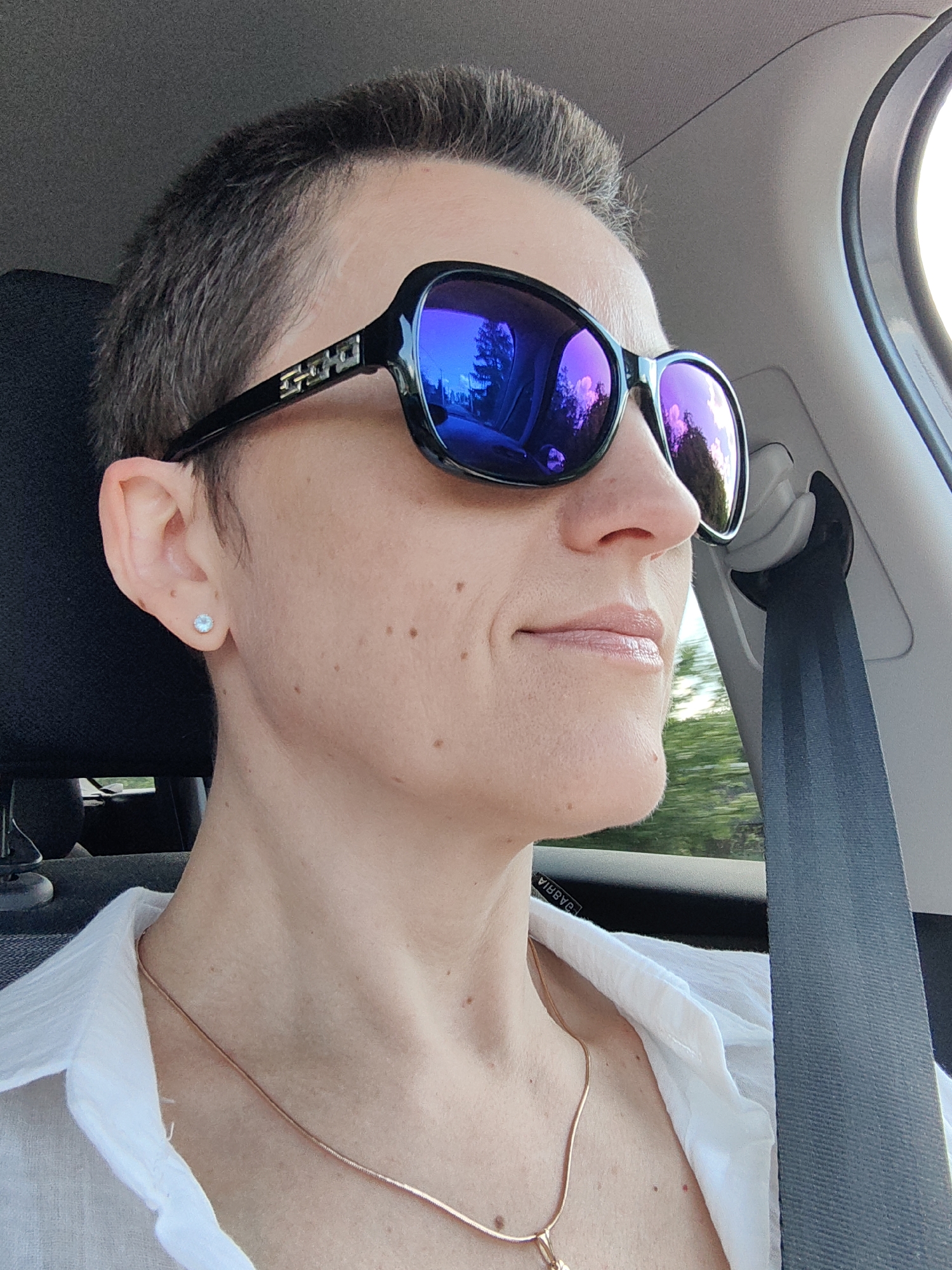 A woman with short, dark hair and sunglasses is pictured from the chest up, looking to the right. She is wearing a white collared shirt and a rose gold necklace. A blue seatbelt is visible to her right. The background shows trees and a bright sky, suggesting the scene takes place outdoors during the daytime, possibly while traveling in a vehicle. The exact location of Ilyanskiy selskiy Sovet, Belarus cannot be determined from the visual information provided.