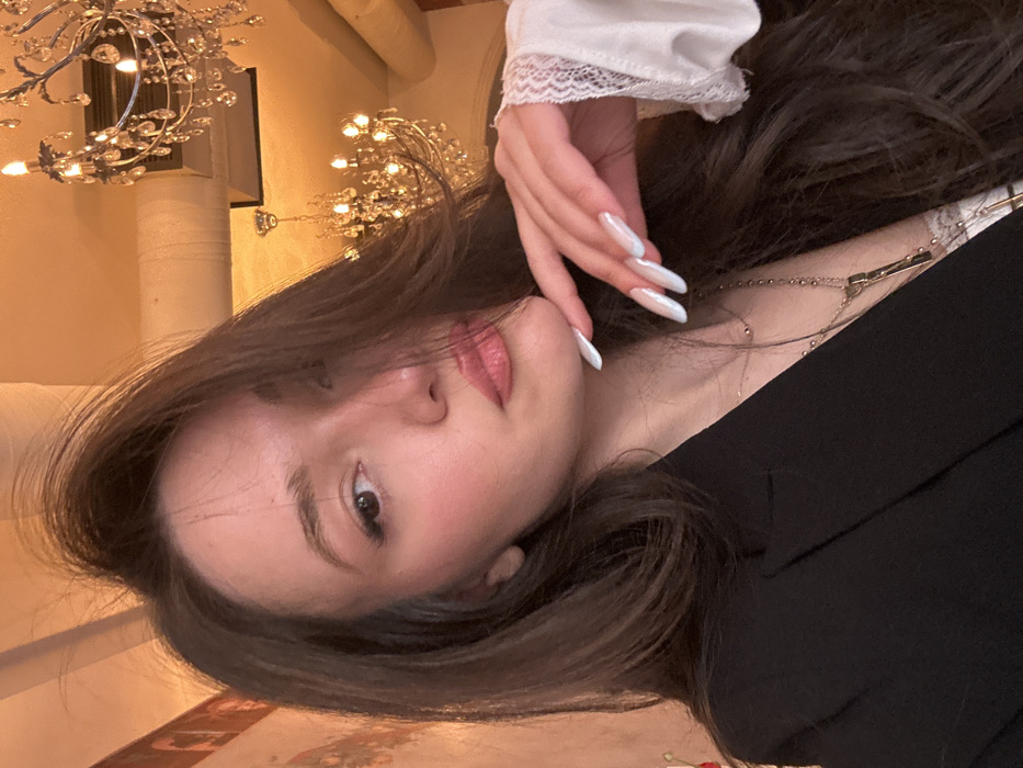 A young adult female is depicted in a close-up, chest-up portrait within an indoor setting in Yerevan, Armenia. She has long, dark brown hair, partly obscuring the left side of her face, and is looking directly at the camera. Her left hand, adorned with long, almond-shaped nails featuring a pearlescent white/light blue polish, is positioned with fingers gently touching beneath her chin. She is wearing a black blazer over a white lace-trimmed top, accessorized with a delicate gold chain necklace and a longer gold necklace featuring a rectangular pendant. The background reveals a warmly lit interior characterized by multiple ornate crystal chandeliers suspended from a light-colored ceiling, which also shows visible utility piping. A textured wall in warm tones is partially visible on the left. The environment suggests an upscale restaurant, cafe, or lounge setting within the city of Yerevan.