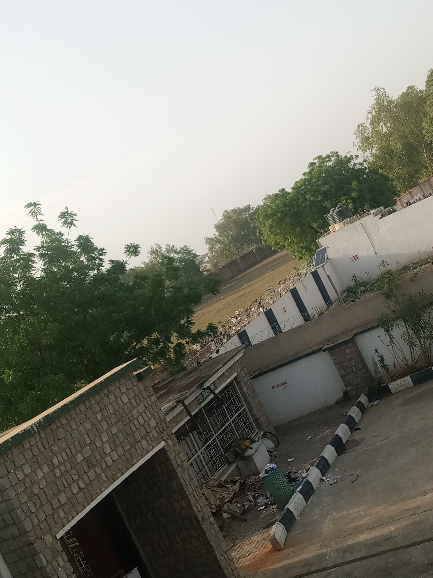 The image shows an outdoor scene in Hotoro North, Nigeria. The foreground features a stone building with a dark opening, and to the right, a paved area with a black and white striped concrete barrier. There is scattered debris, including a green trash bin and loose garbage, on the paved surface. Further back, a white wall with black stripes and barbed wire fencing runs horizontally across the image. A solar panel is mounted on a pole attached to the wall. Behind the wall, a large expanse of dry grass is visible, with several trees scattered throughout. In the background, more trees and a few buildings can be seen under a hazy sky. The overall impression is of an urban or semi-urban environment with signs of upkeep and security measures.