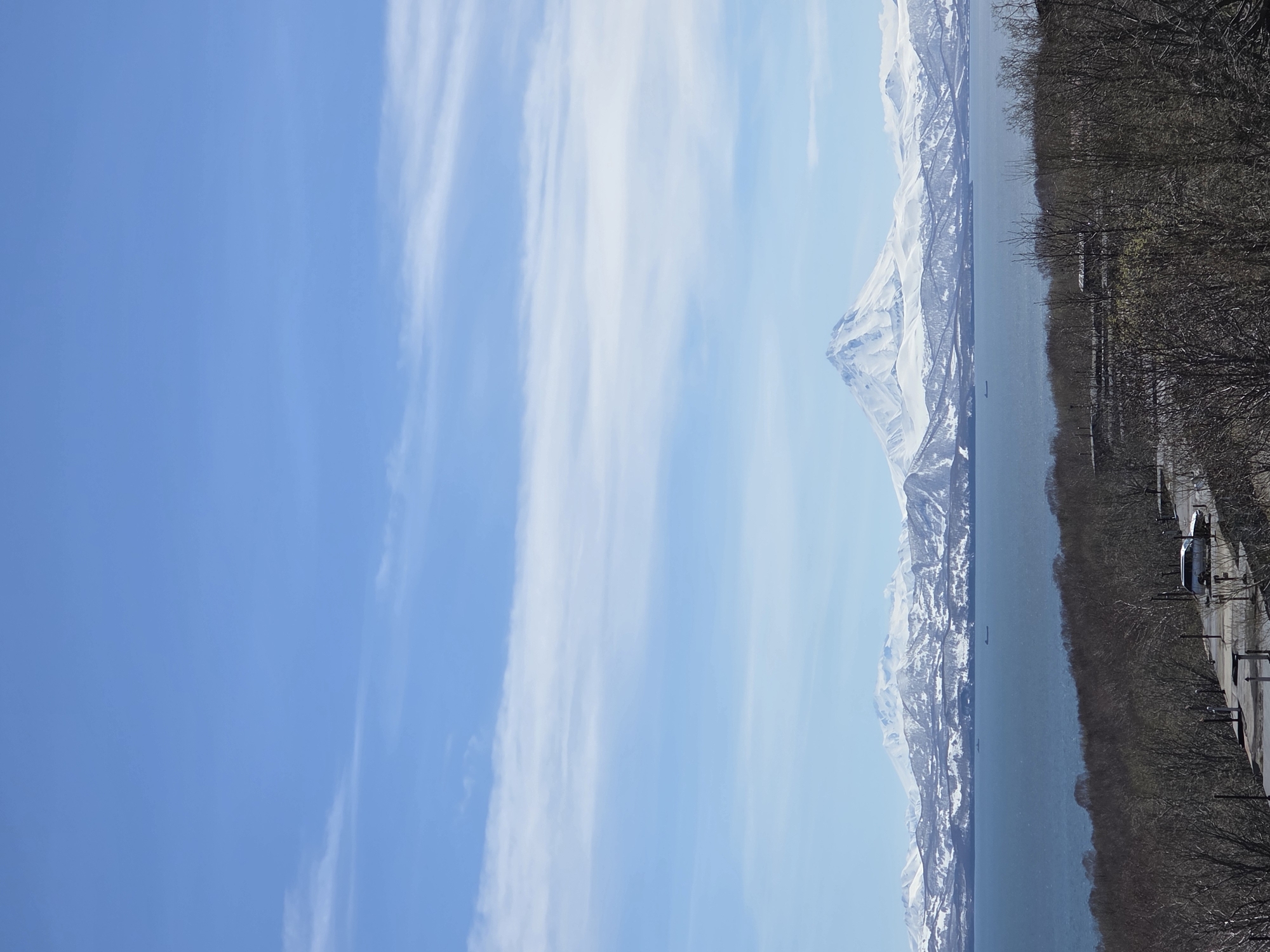 The image shows a mountainous landscape on the right and a blue sky with white clouds on the left. A body of water separates the mountains from the foreground, which is lined with trees and some man-made structures. The location is unknown.