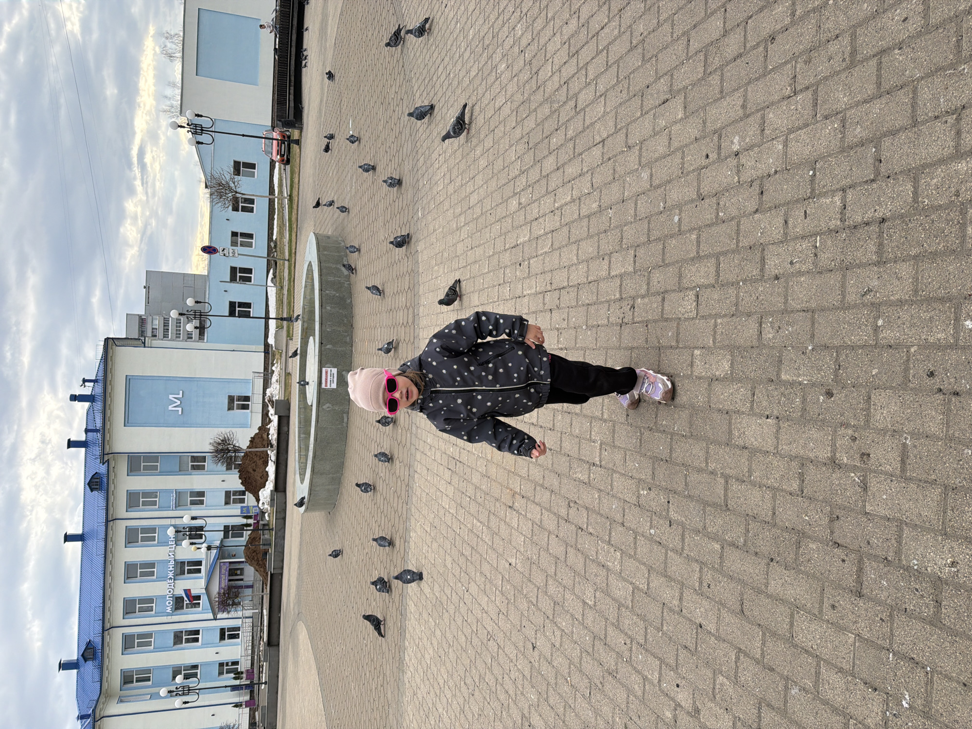 A full-body shot captures a young child standing in an urban paved square, surrounded by numerous pigeons. The child, likely a girl, is positioned slightly off-center in the foreground, facing towards the right, with her head turned towards the camera. She wears a dark jacket with a white pattern (possibly snowflakes), black pants, light-colored sneakers, a light pink beanie, and bright pink framed sunglasses. Her left arm is slightly extended.
The ground is covered in light brown/grey rectangular paving stones, on which many dark-feathered pigeons are scattered, appearing to peck at the ground or move around. To the child's left, a circular concrete structure, possibly the base of a fountain, is visible.
In the background, several buildings line the square under a partly cloudy, bright sky. A prominent light blue and white building features multiple windows and a blue roof. A Russian flag is visible near its entrance. Text on this building reads "МОЛОДЕЖНЫЙ ЦЕНТР" (Molodezhny Tsentr, meaning "Youth Center") and a large "M" is visible on a higher section of the building. Streetlights with multiple globes are also present. Patches of dirty snow are visible near the base of the buildings, suggesting late winter or early spring. A red car is parked further down the street. Near the concrete structure, a white sign with red text reads "ВНИМАНИЕ!" (Vnimaniye!, meaning "Attention!"). The overall scene depicts a common urban activity of feeding or observing pigeons in a public plaza.