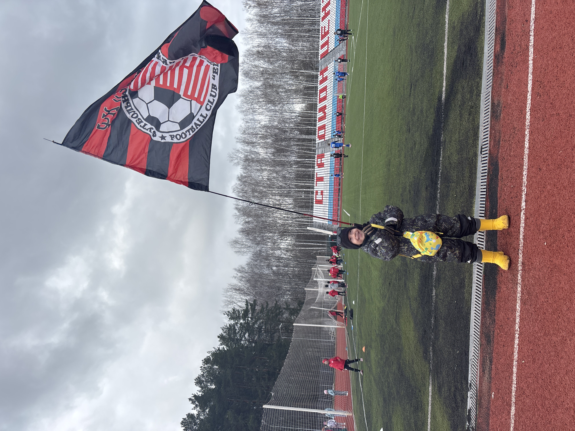 A person is holding a flag at a sports field. The flag is red and black with a soccer ball design and text around it. The person is wearing a dark-colored outfit, yellow boots, and a yellow backpack. The field appears to be a soccer field with a running track around it. In the background, there are trees, bleachers, and other people on the field. The location appears to be Chaikovsky, Russia, as the flag says "FC Chaikovsky". The sky is overcast.