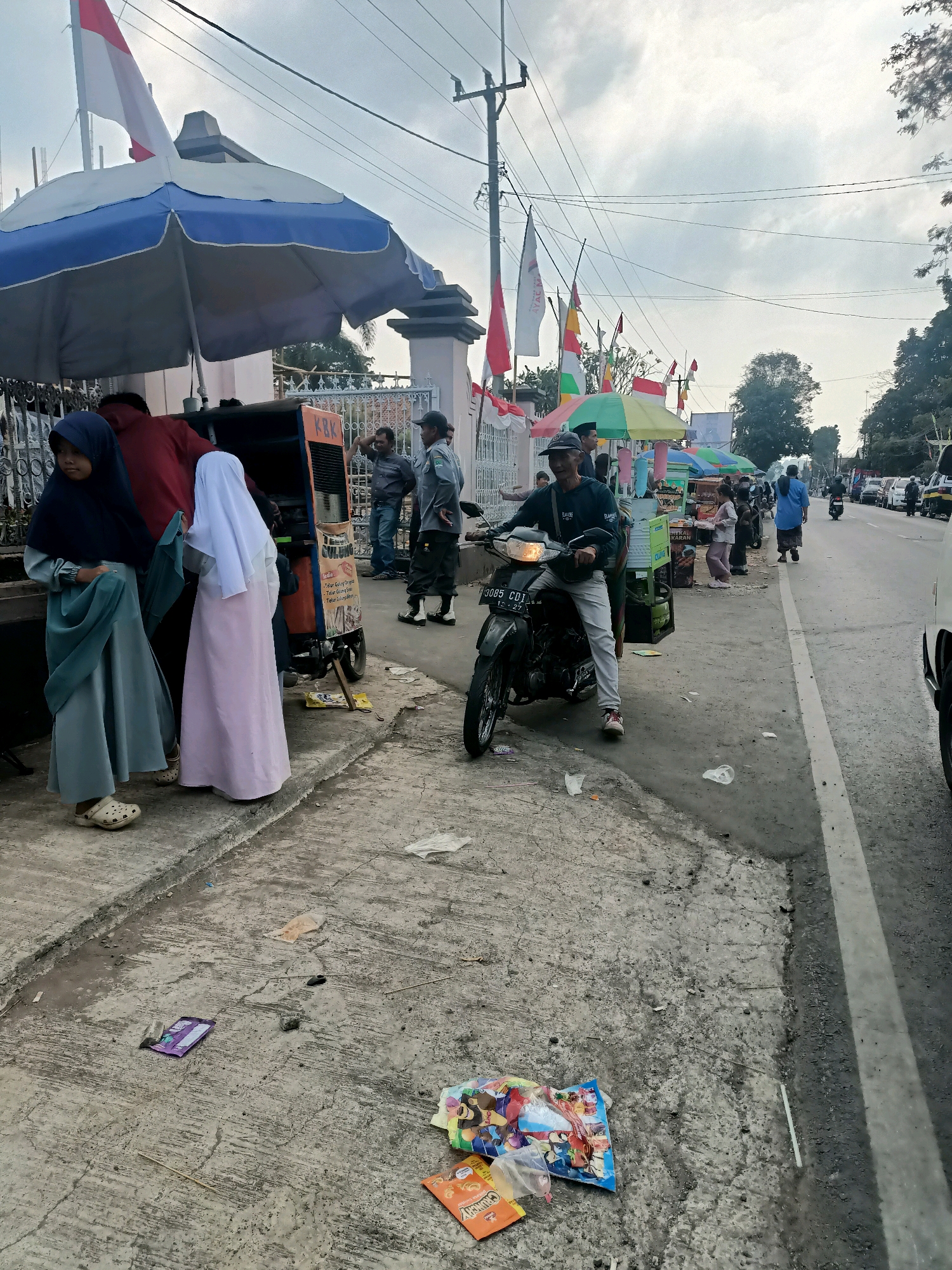 A street scene in Jatipamor, Indonesia, on an overcast day. Two young girls in hijabs stand near a food cart. A man on a motorcycle, wearing a dark jacket and light pants, waits by the cart. Various stalls with colorful umbrellas line the street, and Indonesian flags are visible. In the foreground, scattered wrappers lie on the concrete sidewalk. Cars and other motorcycles are parked or moving along the road. The environment appears to be a busy marketplace or street vendor area.