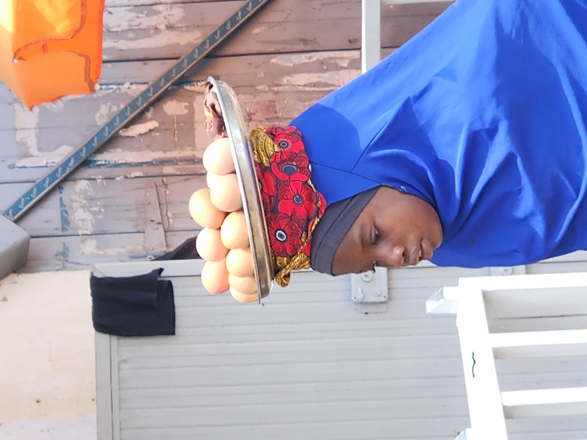 A young person wearing a bright blue hijab and a patterned headscarf carries a metal tray laden with eggs on their head. The eggs are pale brown and arranged in a neat circular pattern. The person's face, in profile, shows them looking downwards with their mouth slightly ajar. Behind them, a rustic wooden door with vertical planks is visible, painted in faded grey. A dark fabric is draped over a wall to the left. The scene suggests an outdoor market or street in a city.